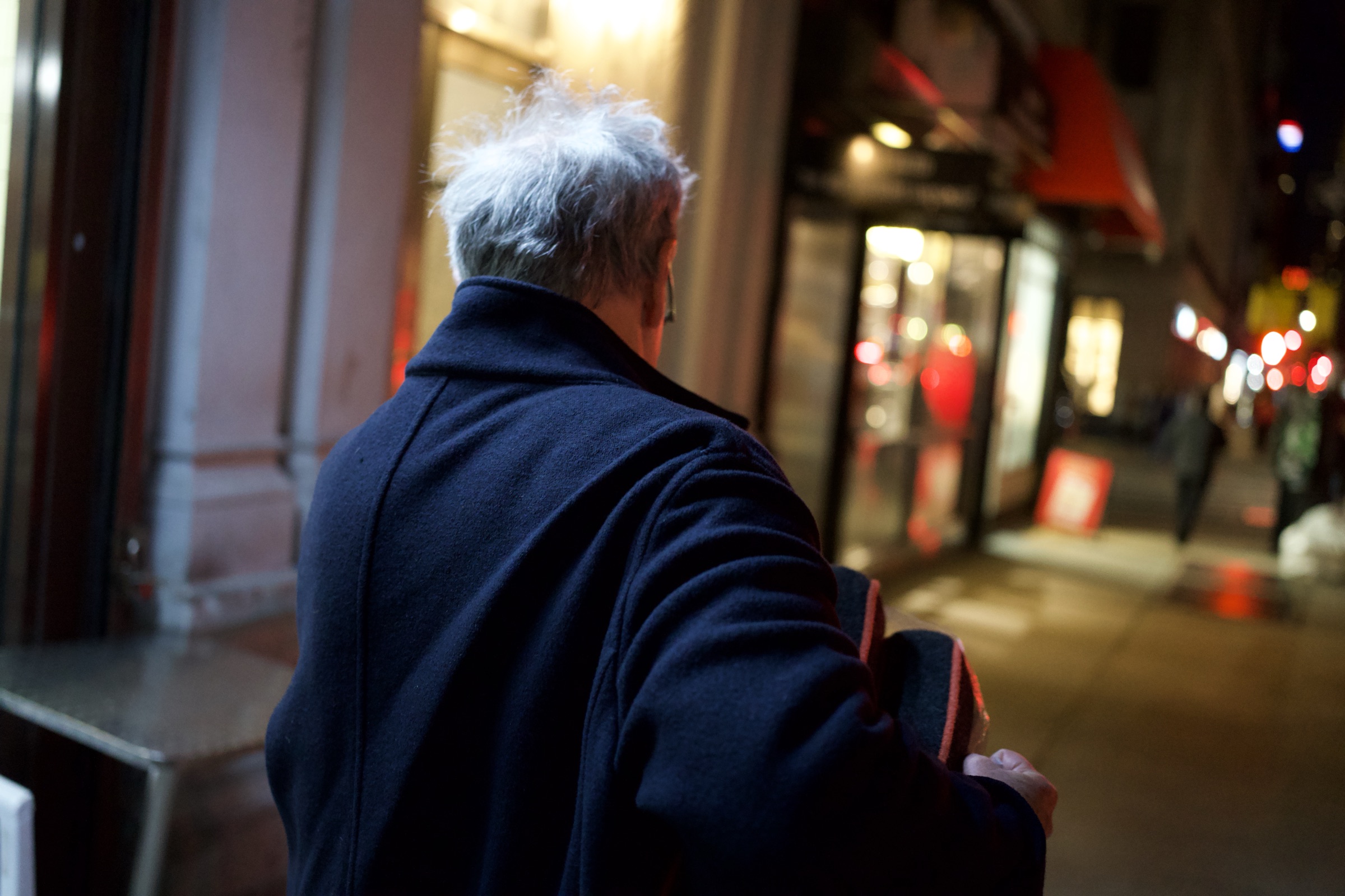 A figure in a dark navy overcoat, silver hair, walking away on a New York City street at night