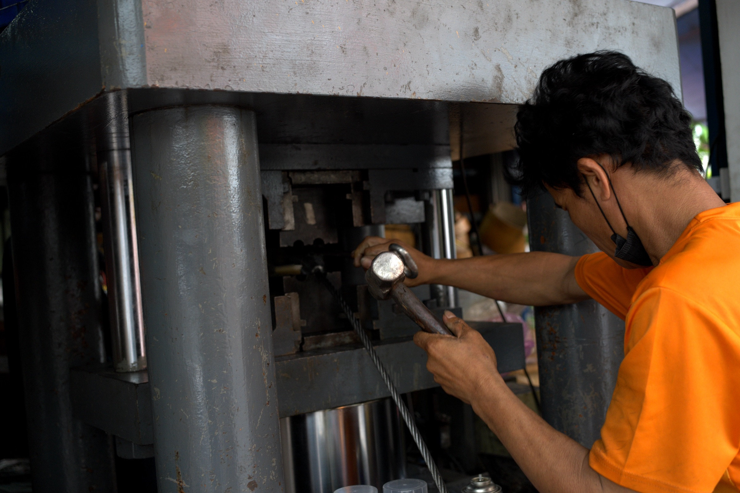 Young metalworker in an orange shirt hammers a piece into an industrial press, face mask pulled down, the machine dwarfing him, natural light from the open shop front