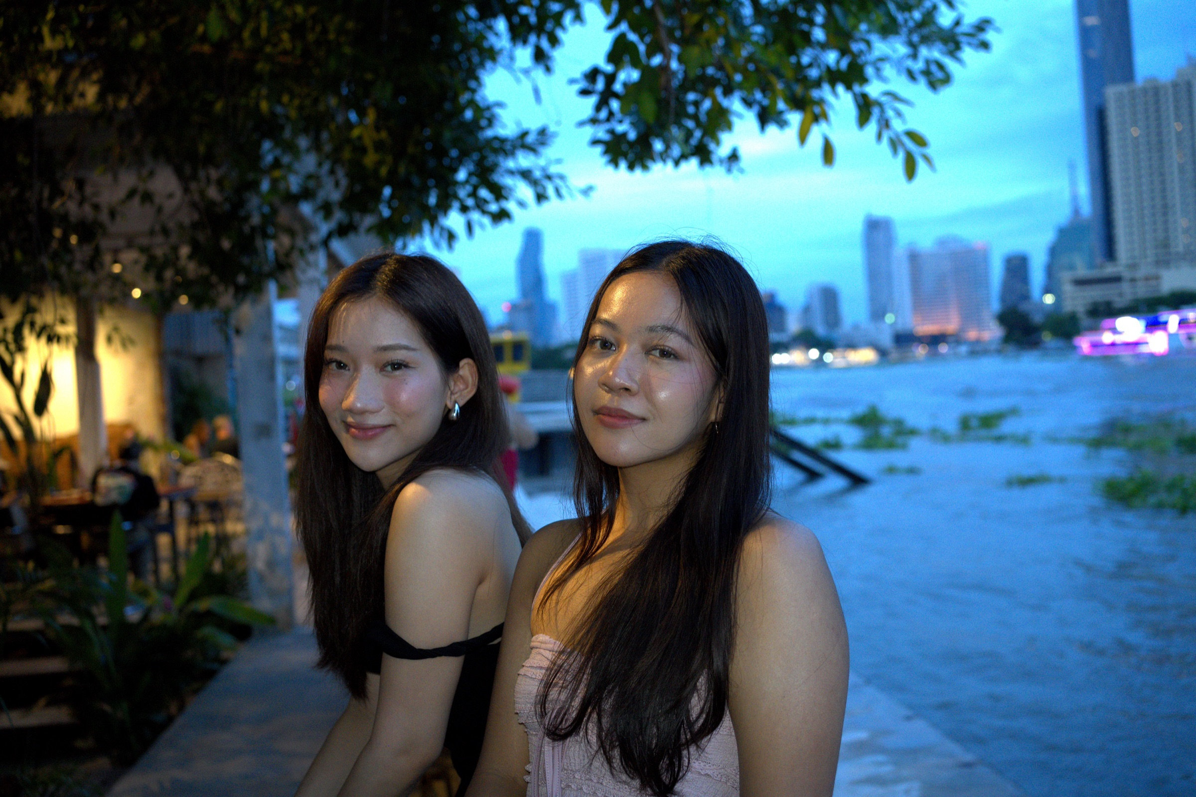 Two young women sit side by side on the Chao Phraya riverfront at blue hour, warm light from a riverside bar behind them, Bangkok skyline rising through the trees across the water