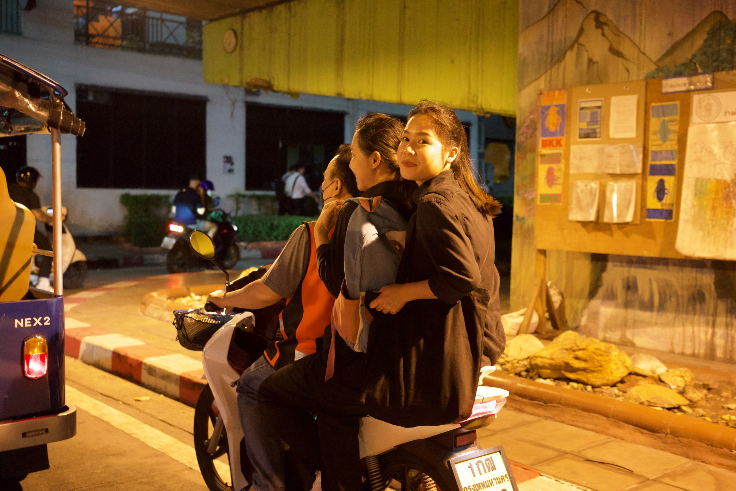 Three people on a motorbike at night in Bangkok, a young woman on the back looking directly at the camera with a subtle smile, BKK signs on a yellow corrugated wall, warm sodium light