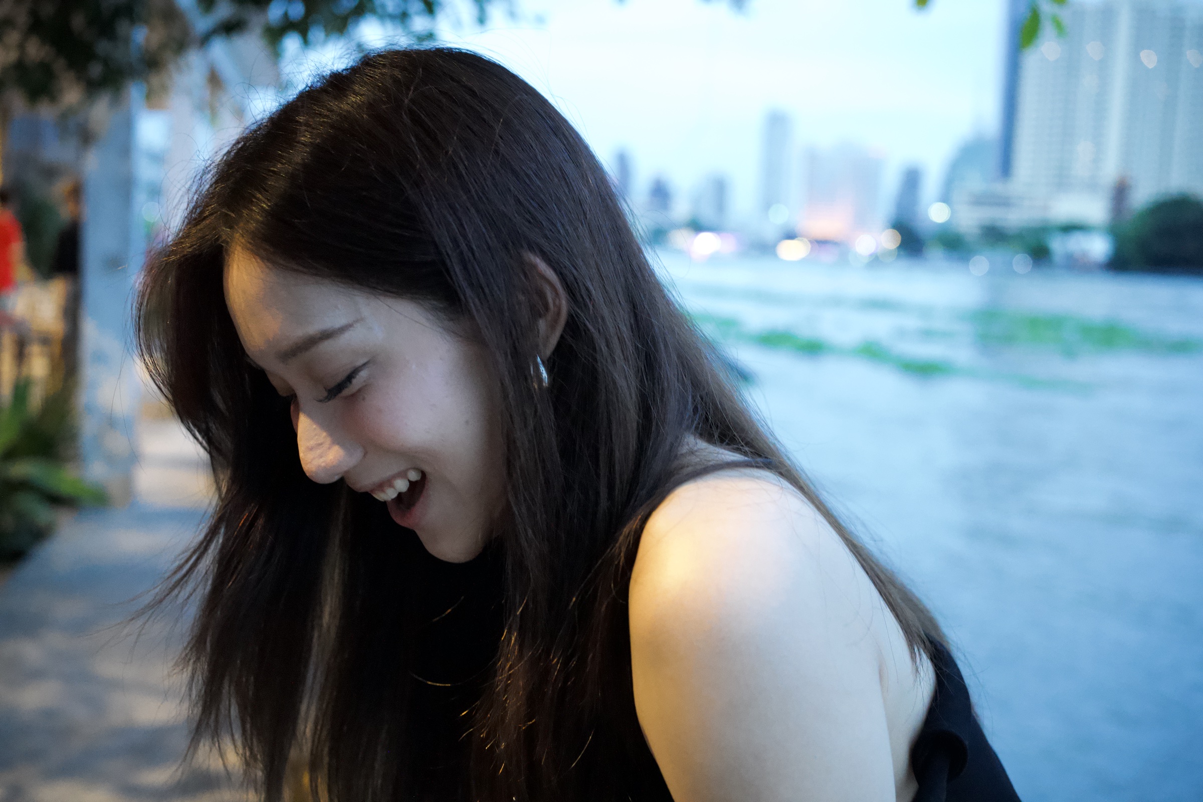 Woman laughing with her head tilted down, hair falling across her face, Bangkok skyline soft behind her at dusk on the Chao Phraya riverfront