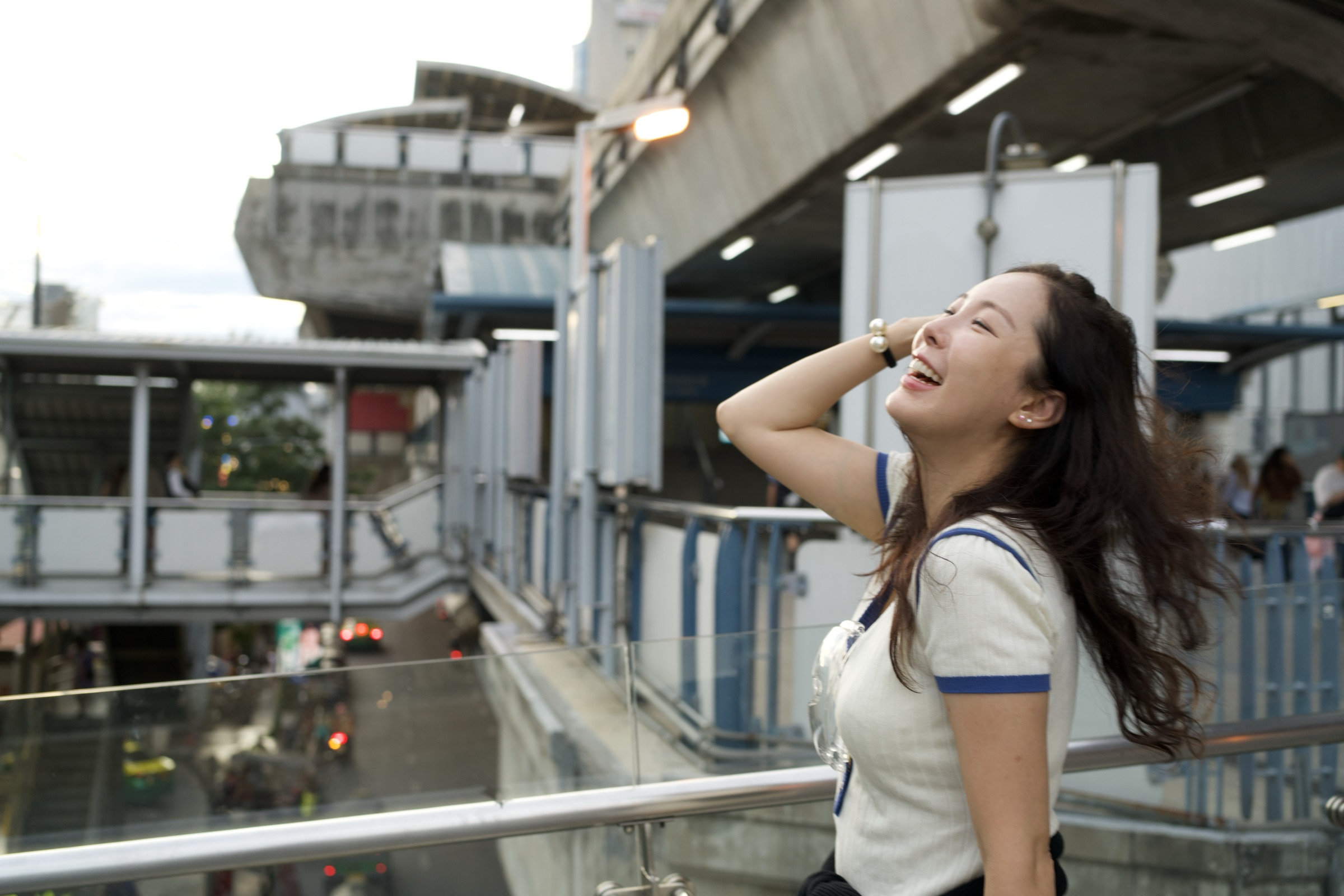 Young woman laughing on a BTS skywalk, head thrown back, hair flowing, white polo and crossbody bag, the brutalist concrete of the Bangkok transit station sharp behind her