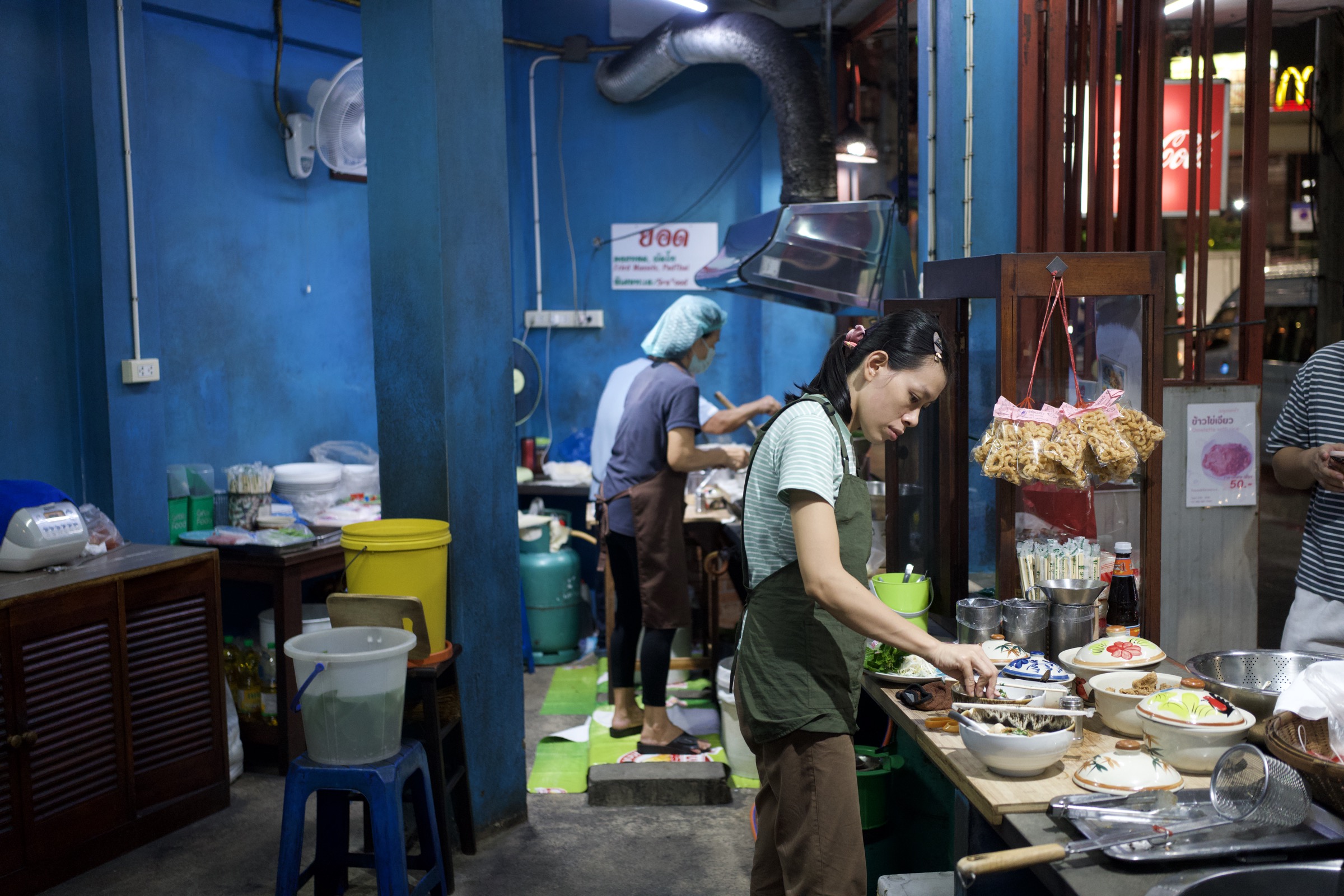Woman in a green shirt prepares food at a blue-painted Bangkok street stall, bags of dried noodles hanging above, another cook working in the vivid blue kitchen behind her