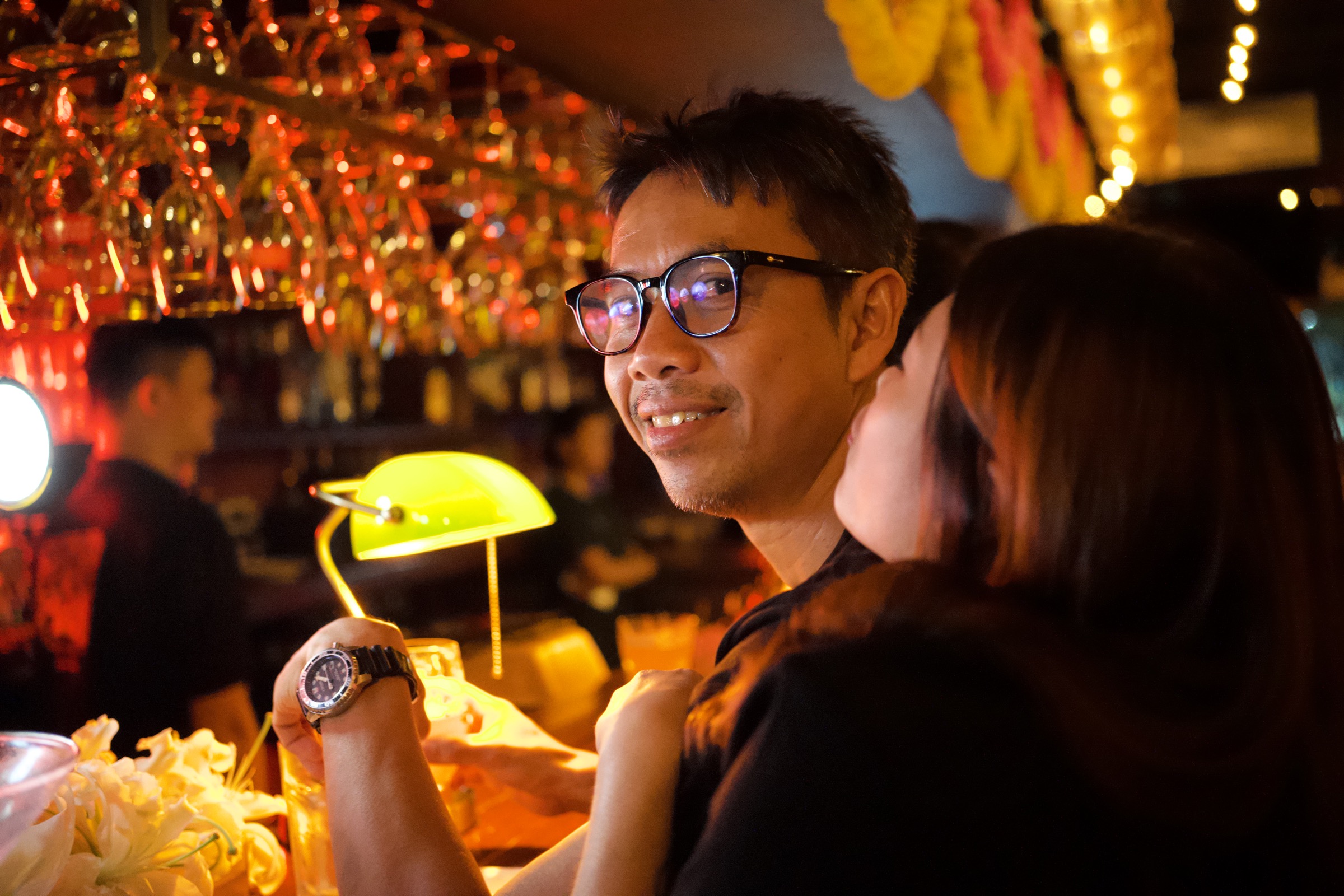 Man with glasses smiles softly as a woman leans close at a Bangkok bar, green banker's lamp and warm lights behind them