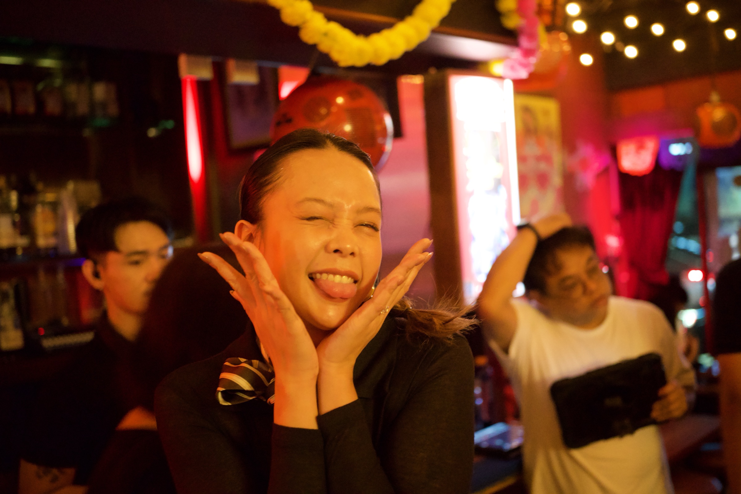 Woman cups her face in her hands and beams at the camera in a Bangkok bar, red and pink neon glow behind her, friends dancing in the background
