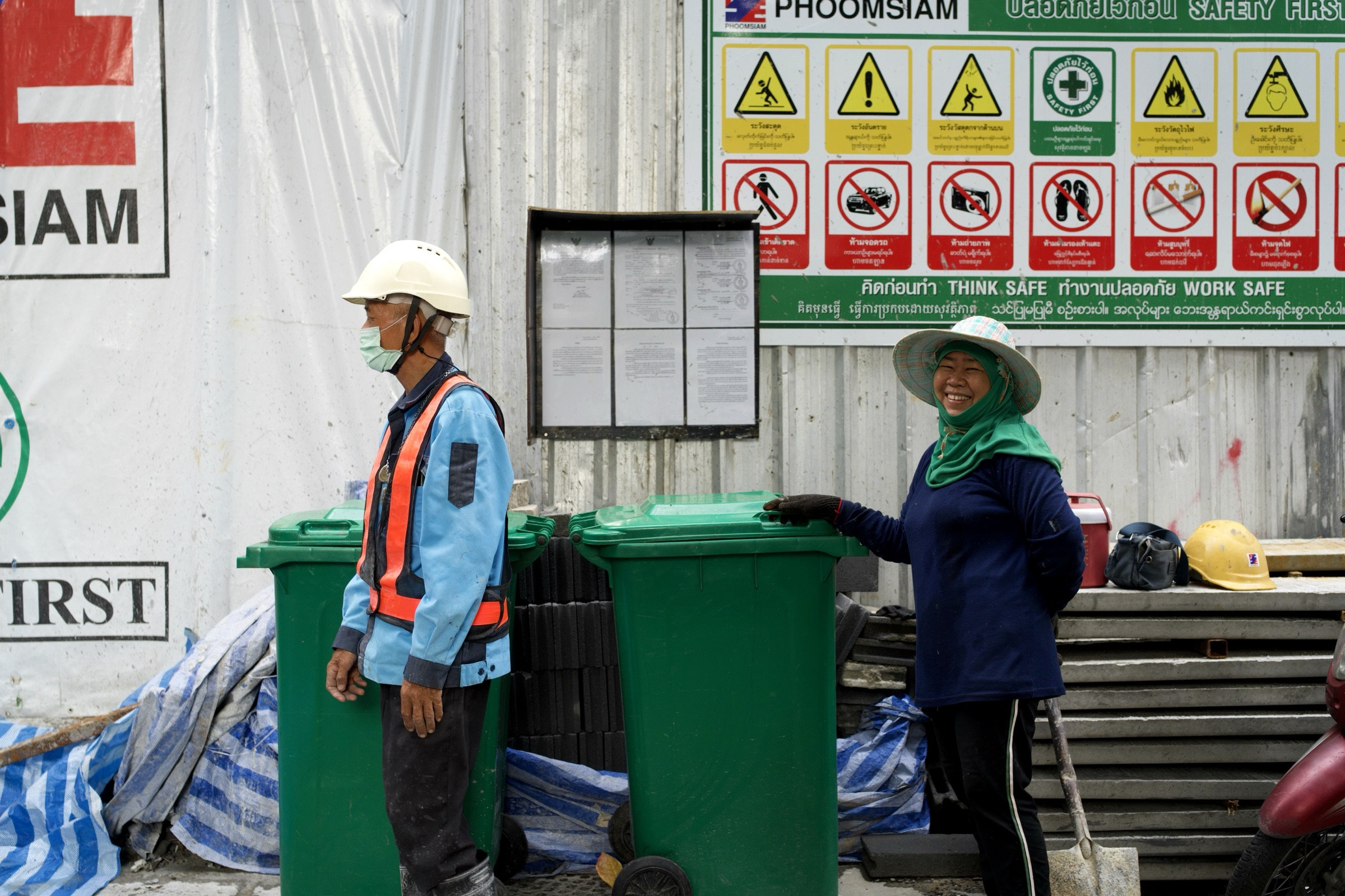 Construction worker in a wide-brimmed hat and green scarf smiles warmly at the camera, safety signs behind her