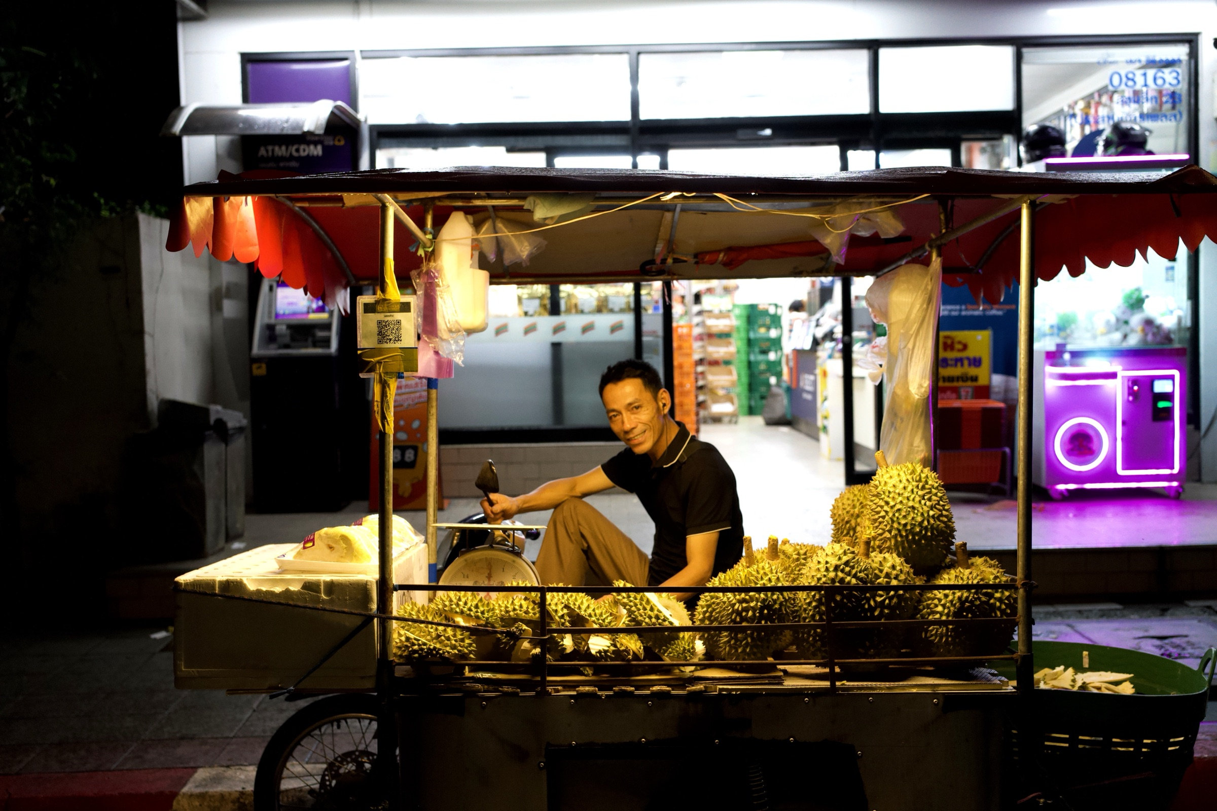 Durian vendor seated in his mobile cart at night, knife in hand, looking directly at the camera, whole durian fruits surrounding him, an ATM and purple-lit vending machine glowing behind