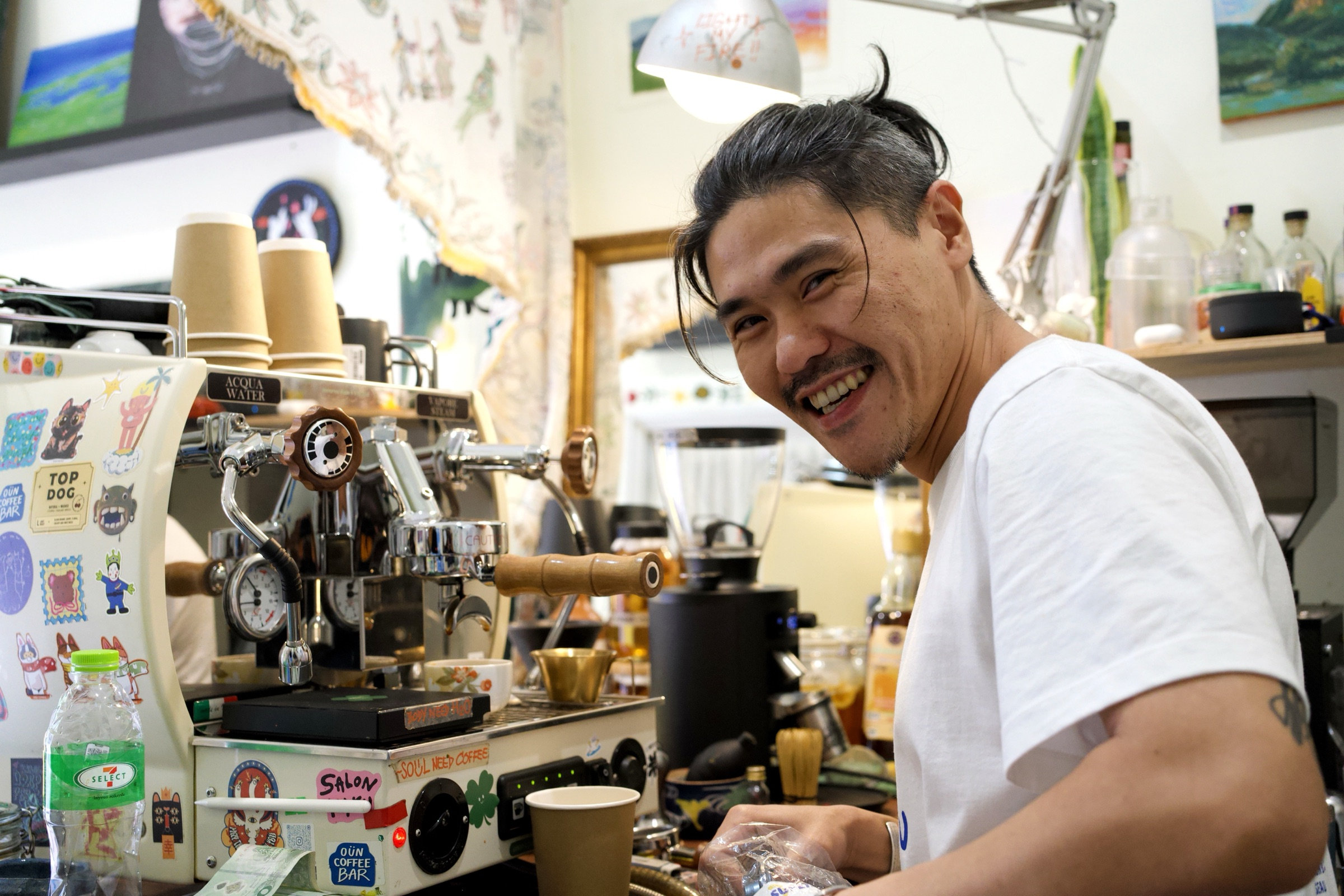 Barista grinning behind a sticker-covered espresso machine at Soul Need Coffee, Bangkok, tattoo on his forearm, paintings on the wall, warm light from an adjustable lamp