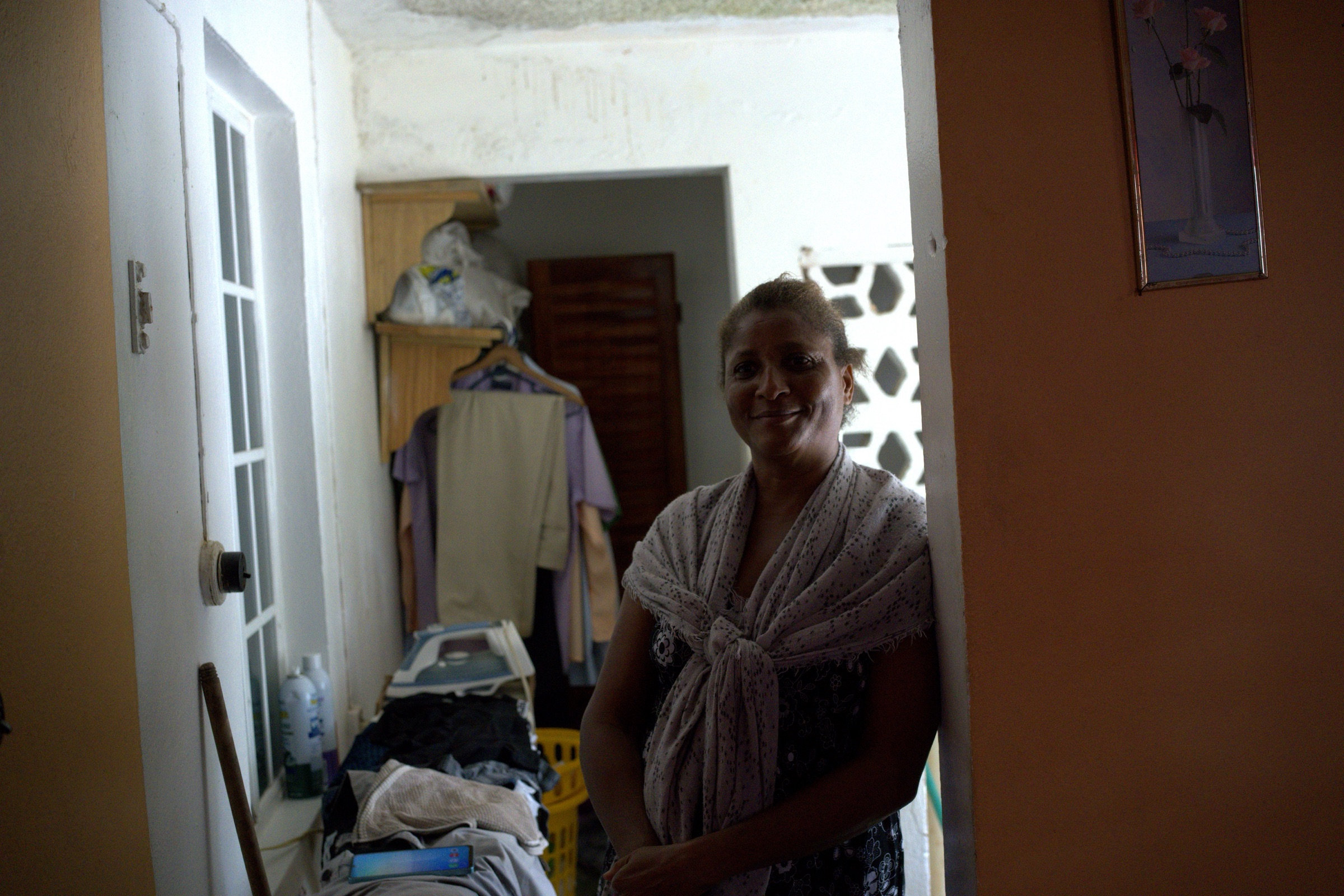 Woman stands smiling in a terracotta-walled doorway, a polka-dot shawl tied at her chest, laundry filling the hallway behind her