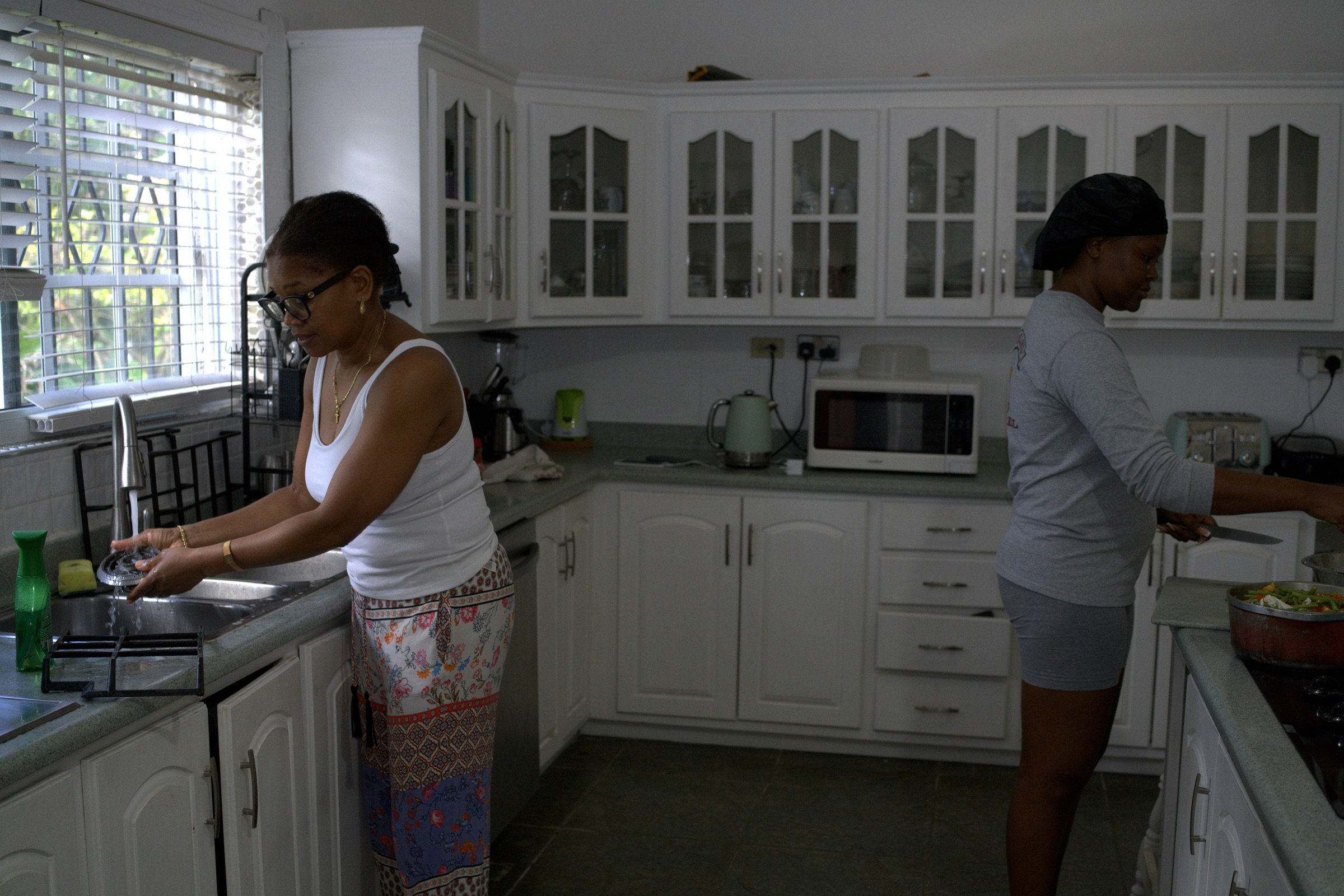 Two women work side by side in a sunlit Jamaican kitchen, one washing dishes, the other chopping at the counter