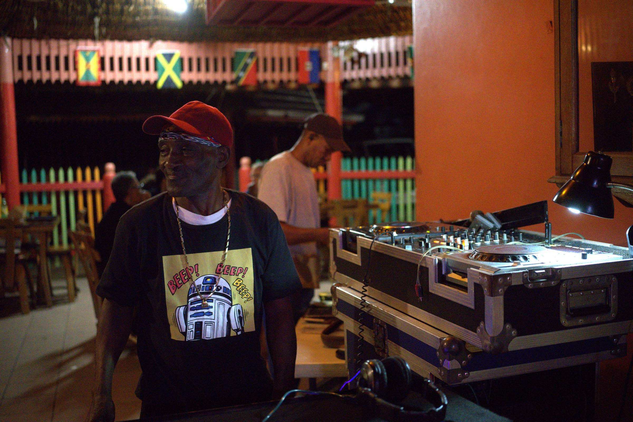 Older man in a red cap and an R2-D2 Beep Beep t-shirt stands near a DJ setup, Jamaican flags strung across the wooden railing behind him, the dance hall warm and dark beyond