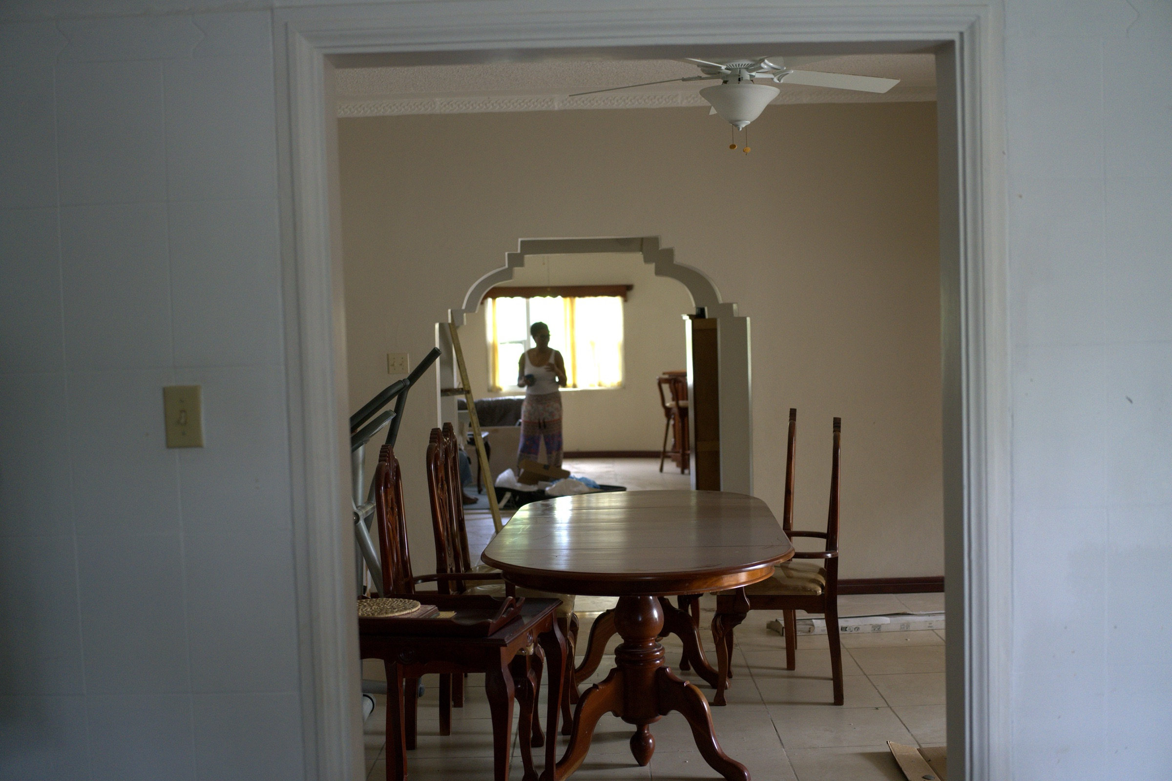 View through a doorway into a dining room, dark wood table and chairs in the foreground, a ceiling fan overhead, and through a decorative archway beyond, a woman silhouetted against a yellow-curtained window deep inside the house