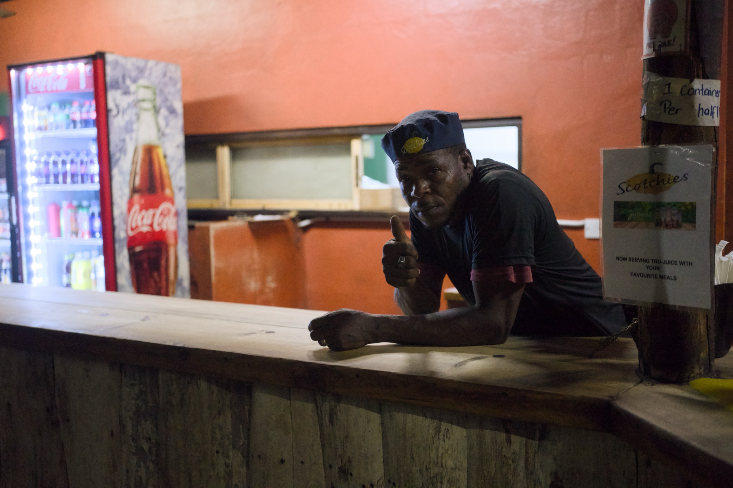 Man in a Scotchies cap leans across the wooden bar counter and points directly at the camera, orange wall and a Coca-Cola fridge glowing behind him
