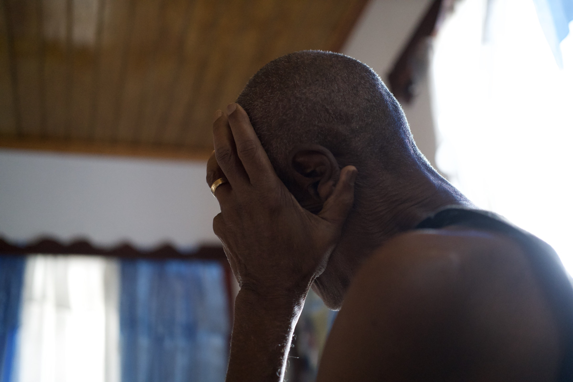 Man sits with his back to the camera, hand pressed against his head, gold ring catching the light, wooden ceiling above, blue curtains and a window glowing behind him