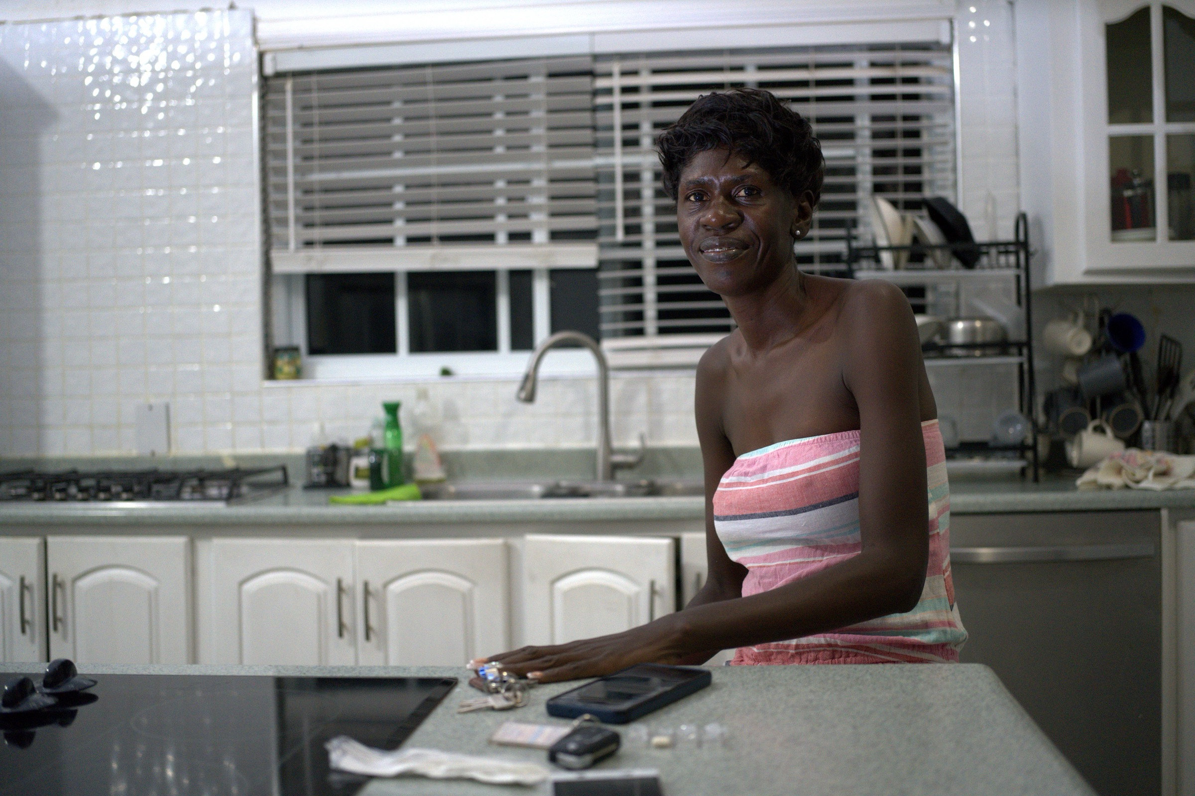 Woman leans on a kitchen counter at night, looking directly at the camera with quiet warmth, keys and phone beside her hand, blinds and white cabinets behind her