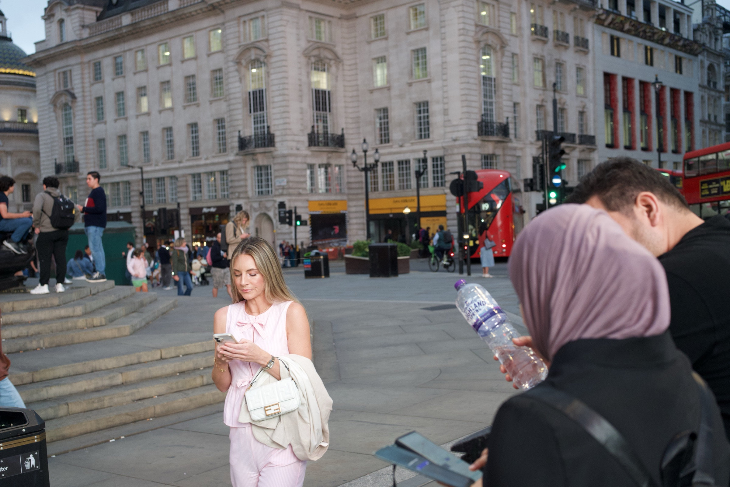 Woman in a pink sleeveless outfit looks down at her phone at Piccadilly Circus, while another in a pink hijab and black jacket holds a water bottle beside her, red double-decker bus and Georgian facades behind
