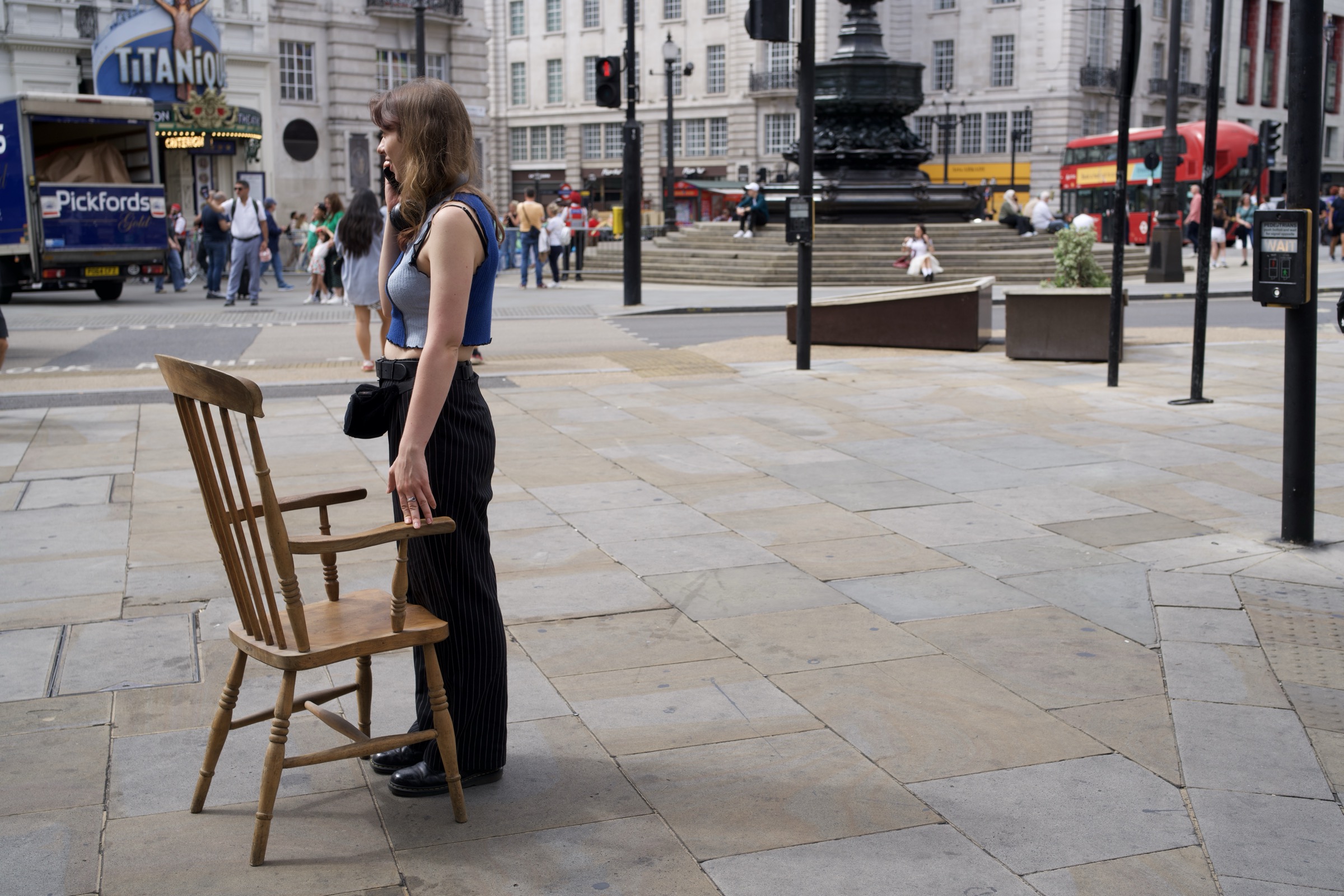 Woman stands beside a wooden chair in the middle of Piccadilly Circus in broad daylight, Titaniq signage and Pickfords truck behind her, the open square stretching beyond