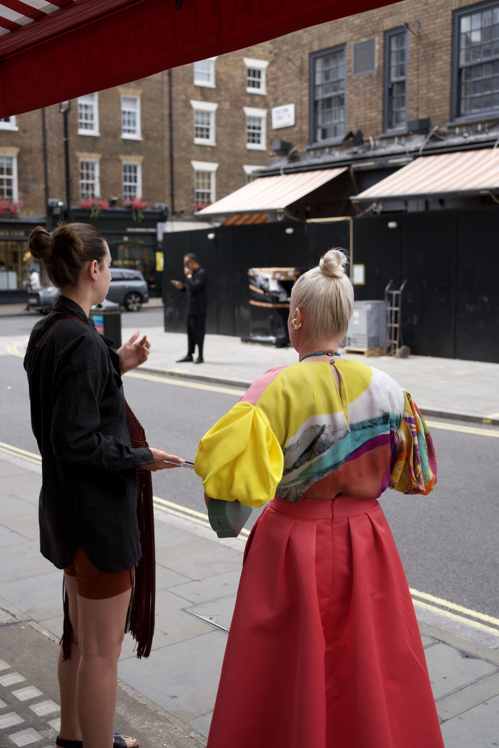 Two women seen from behind on a London side street, one in a multicolor print bolero and full red pleated skirt with a blonde bun, the other in a dark jacket and tan shorts, sunlit brick terraces and a red awning above them