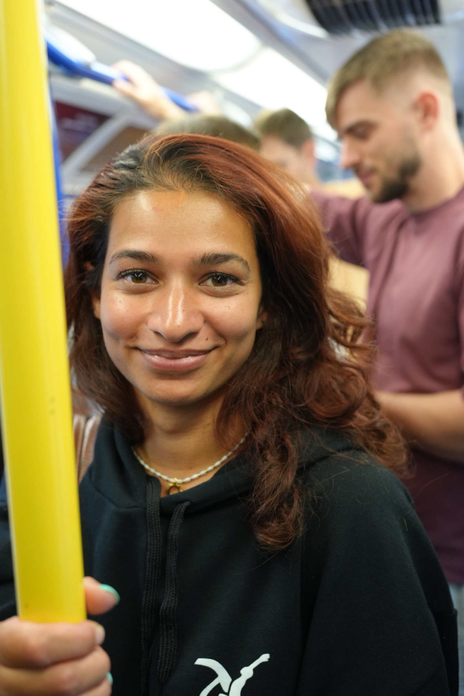 Young woman with auburn hair smiles warmly at the camera on a London bus, yellow handrail cutting across the top of the frame