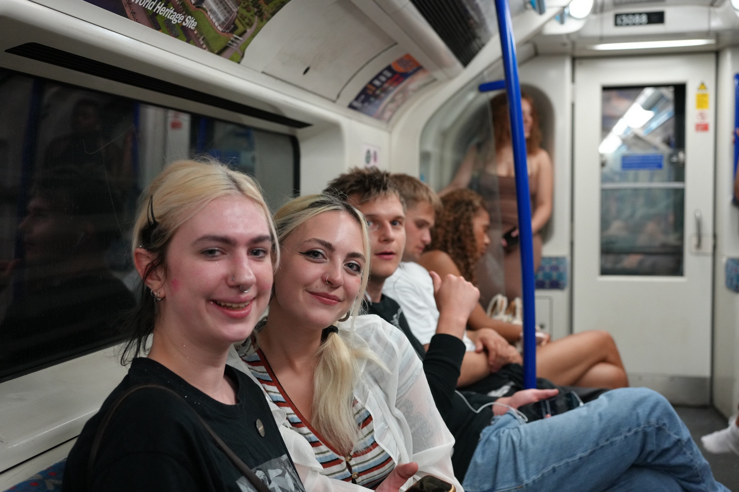 Two young women on the London Underground look directly into the camera with relaxed smiles, friends receding down the row of seats