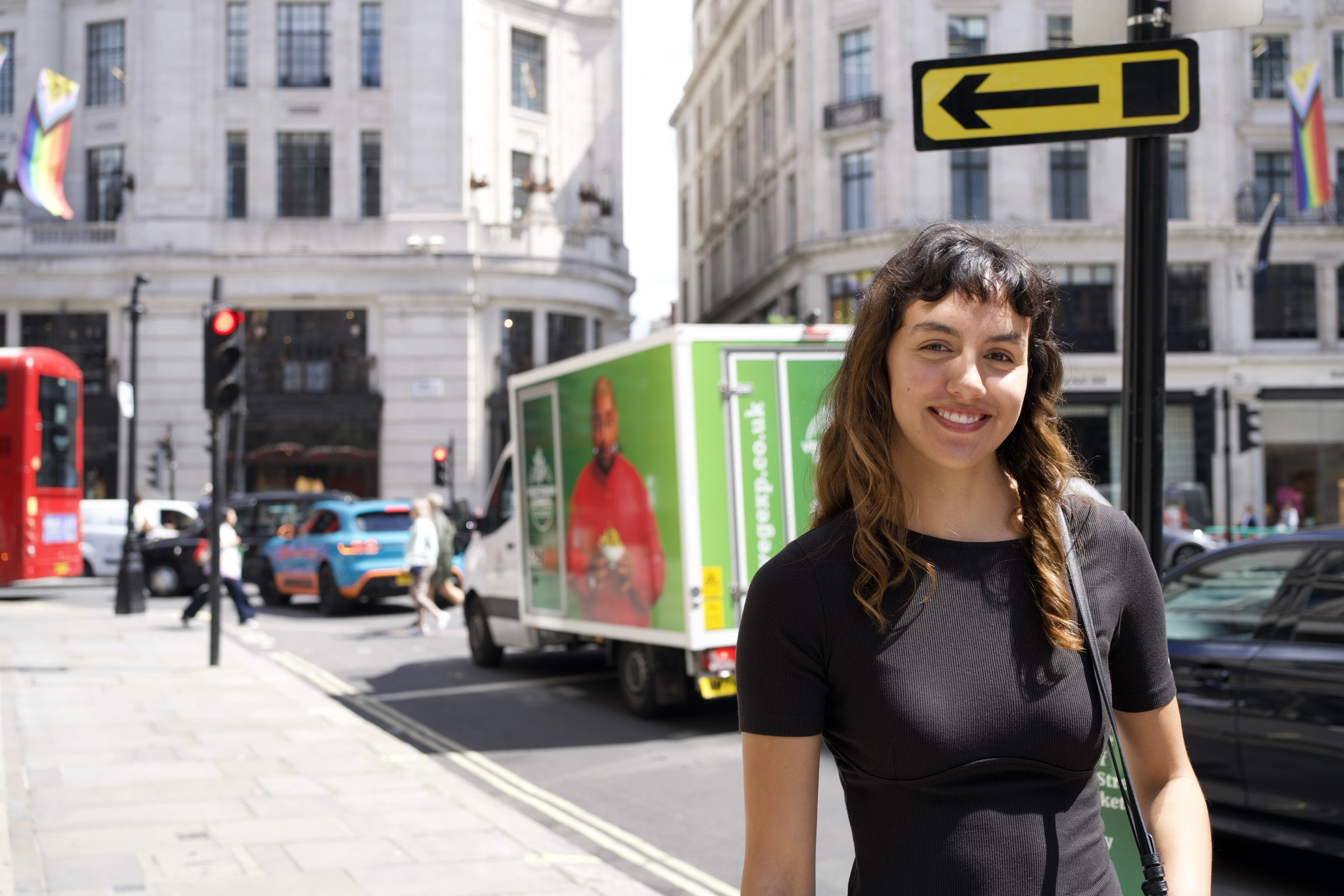 Woman with dark curly hair smiles brightly on a sunny London street, a green grocery van and red double-decker bus behind her, rainbow Pride flags hanging from the stone buildings