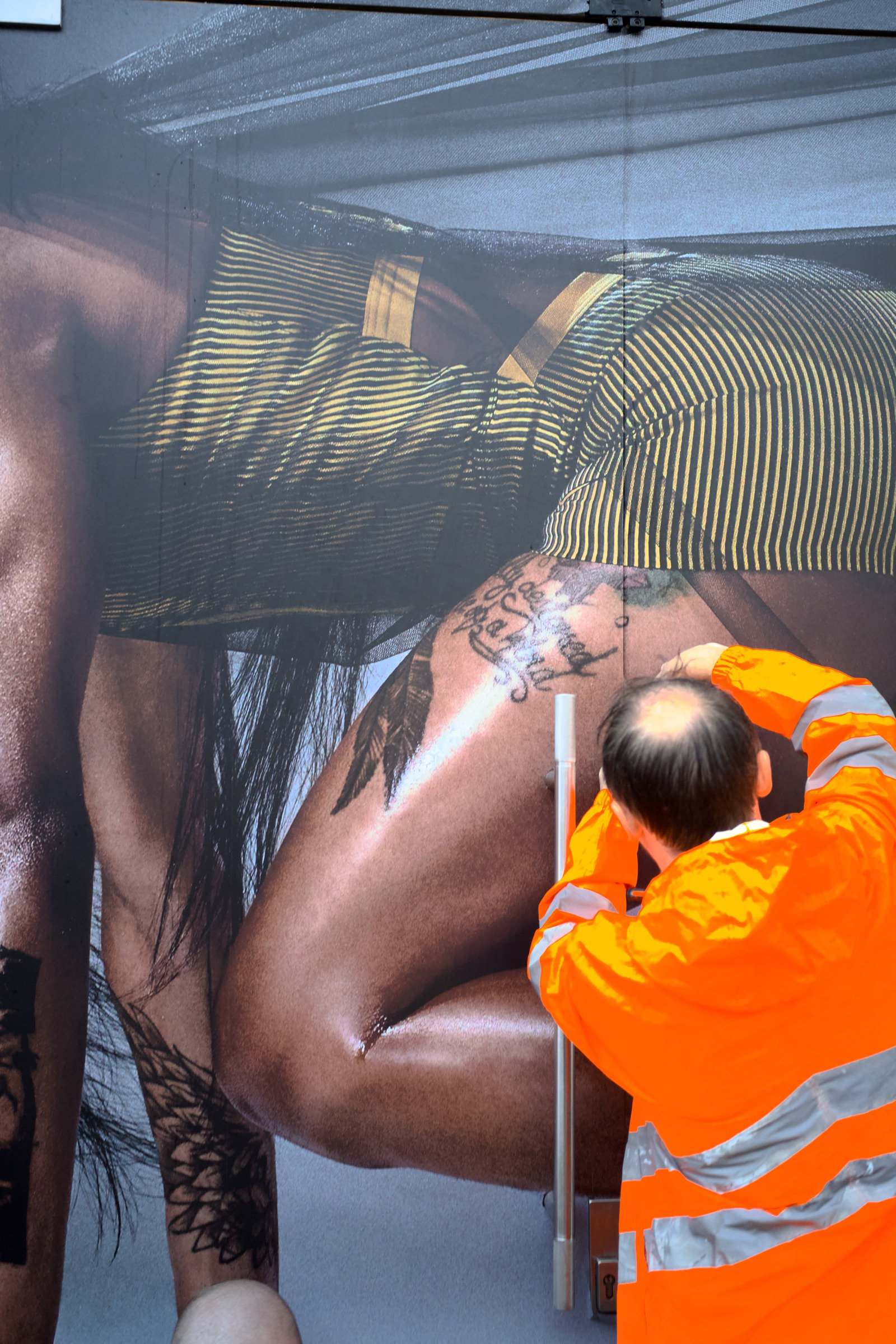 Construction worker in a hi-vis orange jacket kneels at a scaffolding base beneath a towering billboard, the advertisement filling the frame above him, grey London sky beyond