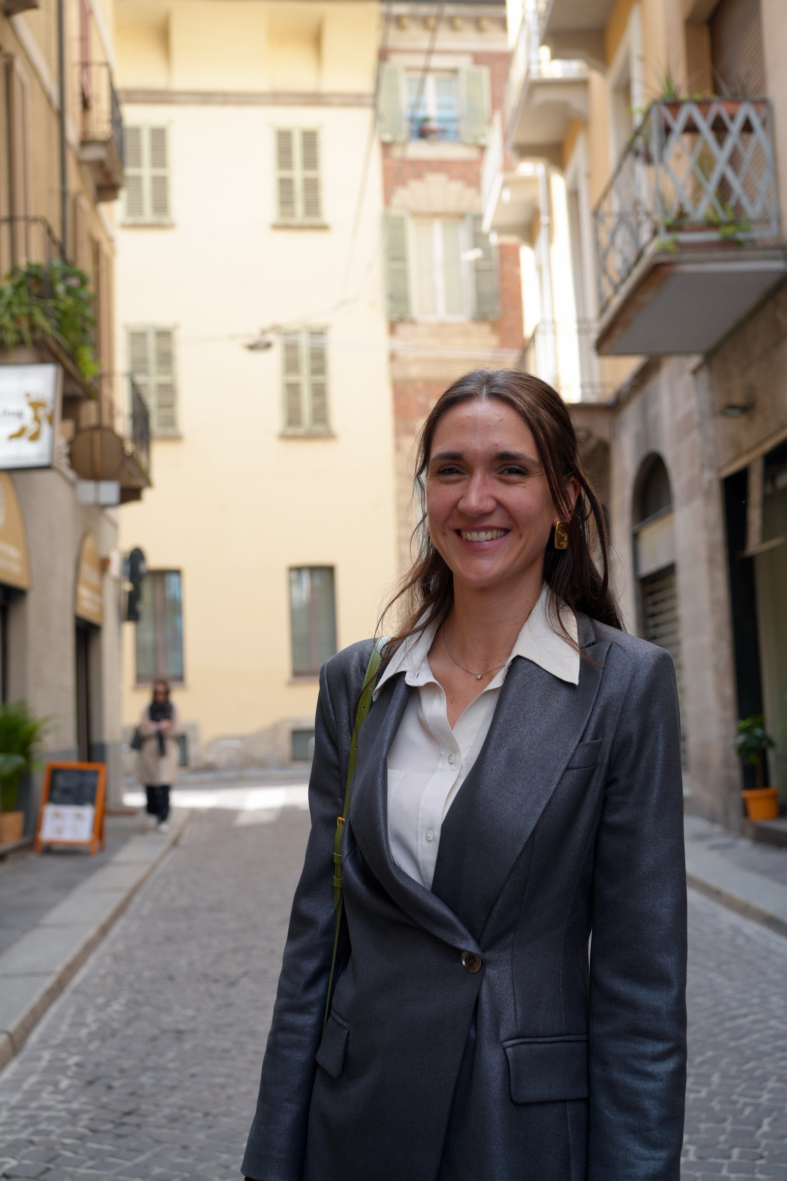 Young woman in a grey blazer and white blouse smiles warmly at the camera on a narrow Italian cobblestone street, golden earrings catching the light, pastel buildings receding behind her