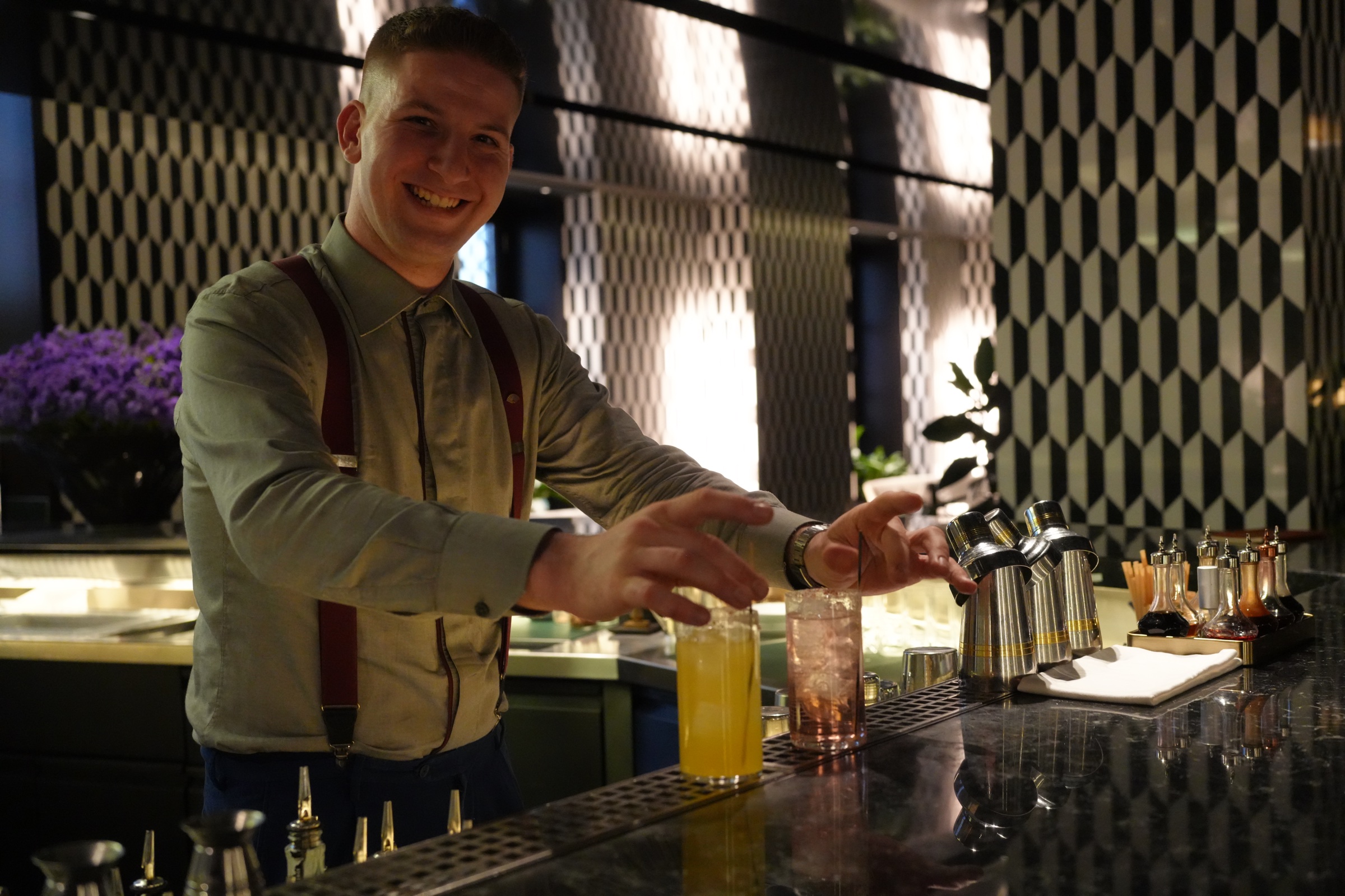 Bartender in dark suspenders and rolled sleeves leans against the bar, bottles lined up behind him, warm amber light from pendant lamps