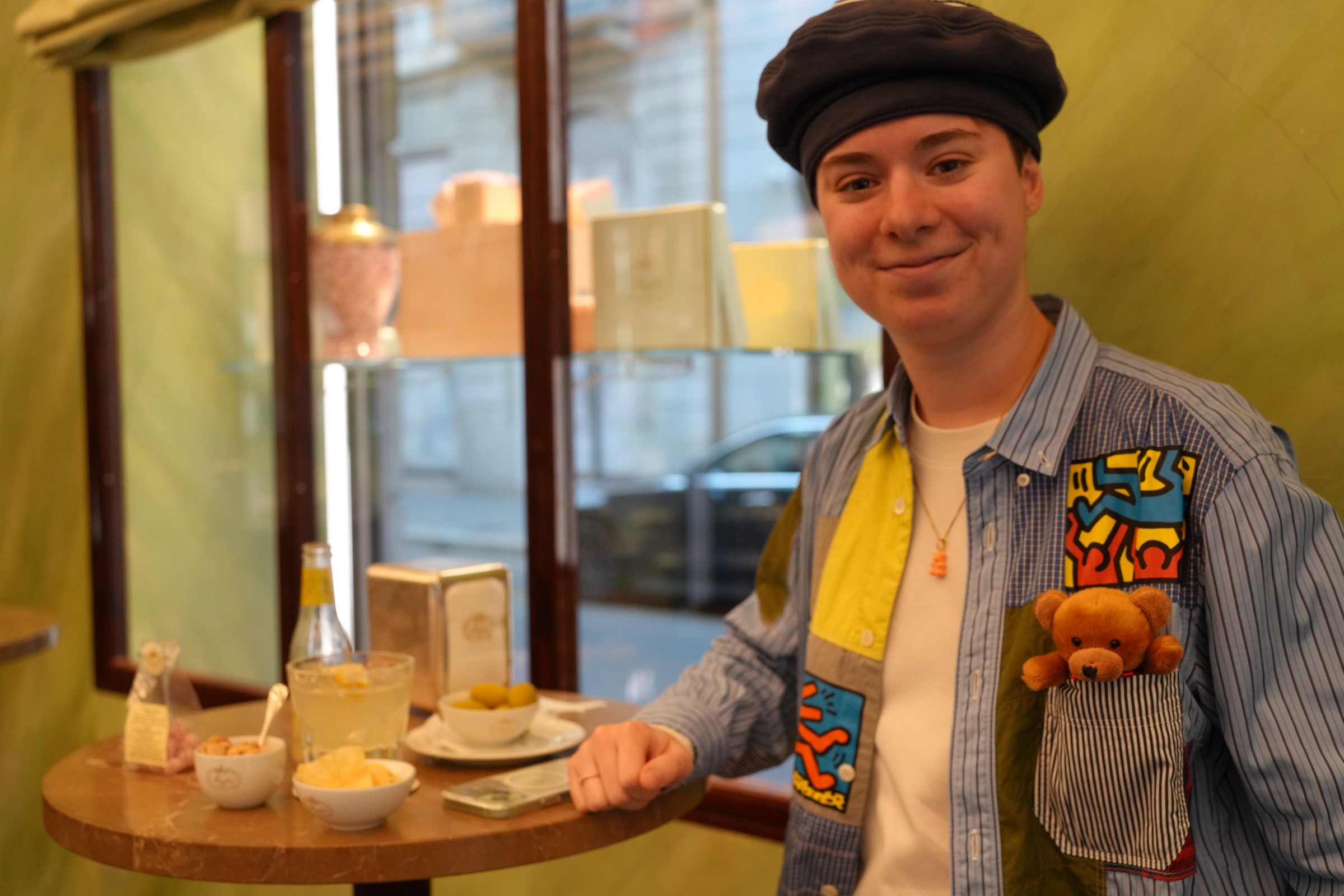 Person in a dark beret and patchwork Keith Haring shirt smiles at an aperitivo table, teddy bear tucked in their pocket, small bowls of olives and chips beside a drink on the round cafe table