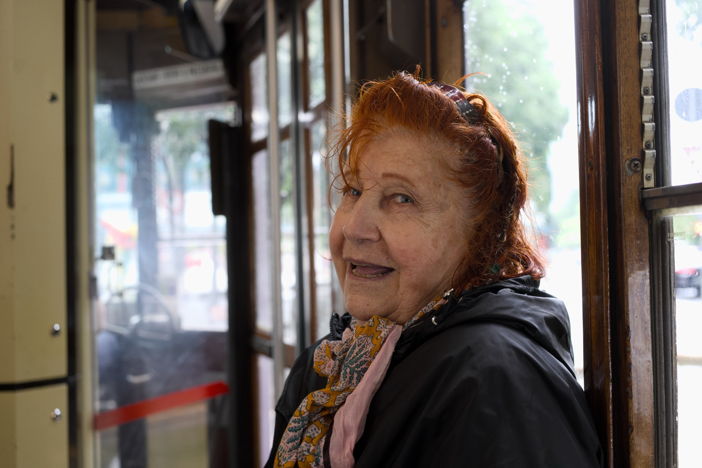Older woman with auburn hair on a Milan tram, looking out the window with a warm, gentle smile, natural daylight catching her face, wooden tram interior behind her