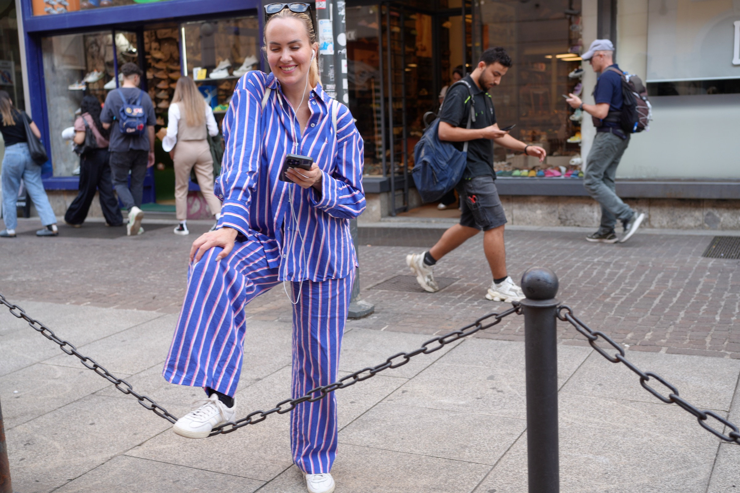 Young woman in a bold blue-and-white striped suit and white sneakers smiles while perching on a chain bollard on a Milan shopping street