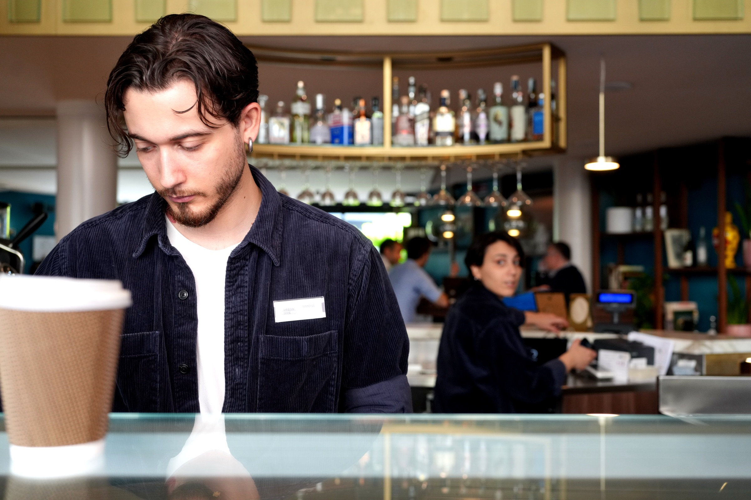 Young barista in a dark corduroy shirt looks down at the counter, a takeaway coffee cup beside him, dark wood paneling and brass fixtures catching the lamplight, a colleague visible in the background
