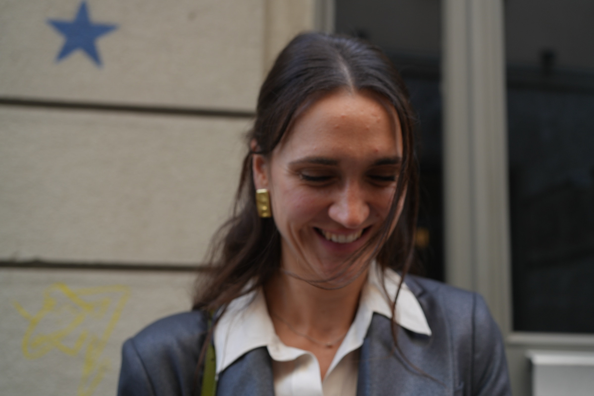 Woman in a grey blazer and gold earring smiles softly downward, caught in a private moment on a Milan street, blue star on the wall behind her