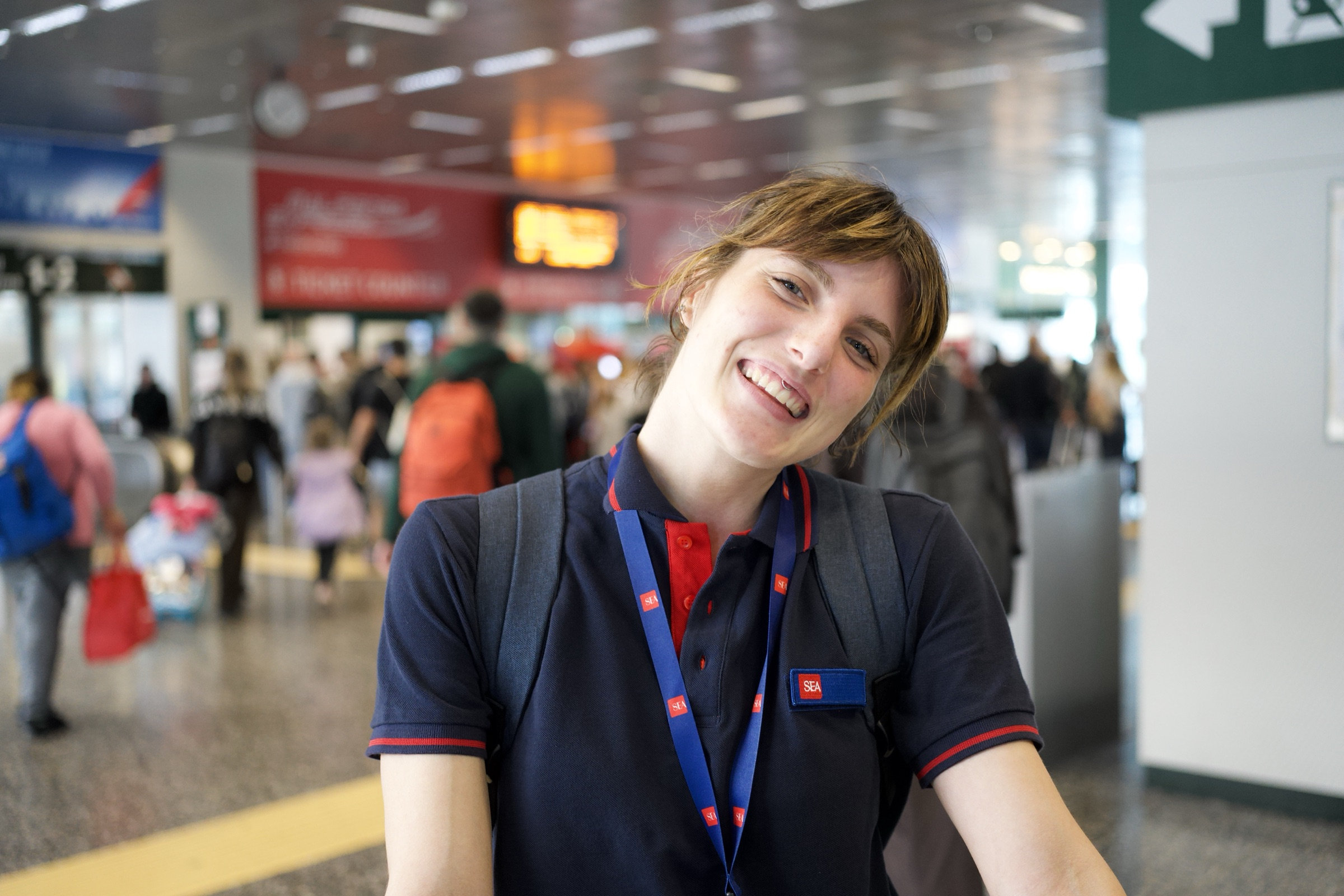 Train station attendant in a navy-and-red polo smiles warmly, bangs swept across her forehead, lanyard around her neck, the bustle of Milano Centrale a blur of travelers behind her