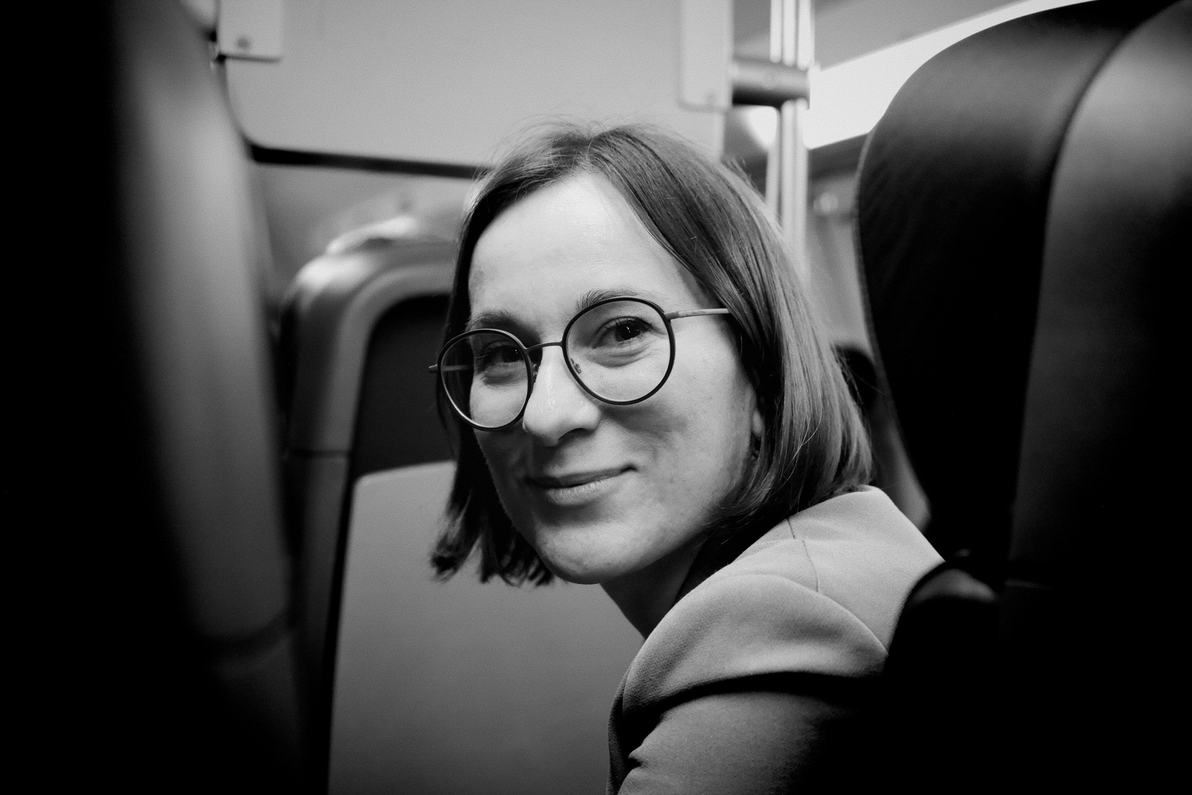 Woman with round glasses and a gentle half-smile sits in a train seat, black and white, soft window light falling across her face