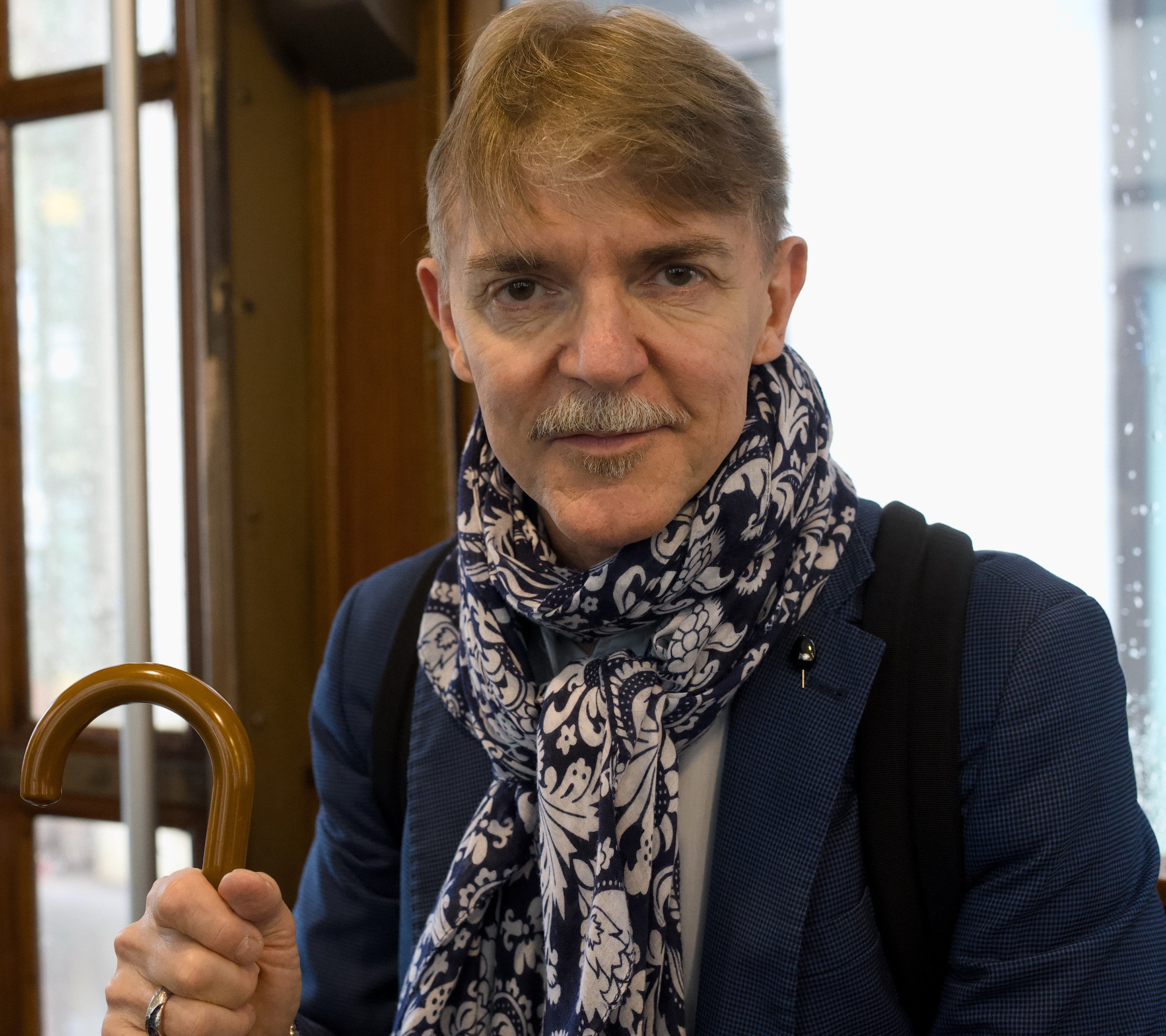 Silver-haired gentleman in a navy blazer and ornate paisley scarf sits on a Milan tram, gazing directly at the camera with quiet dignity, wooden handrail and tram interior behind him