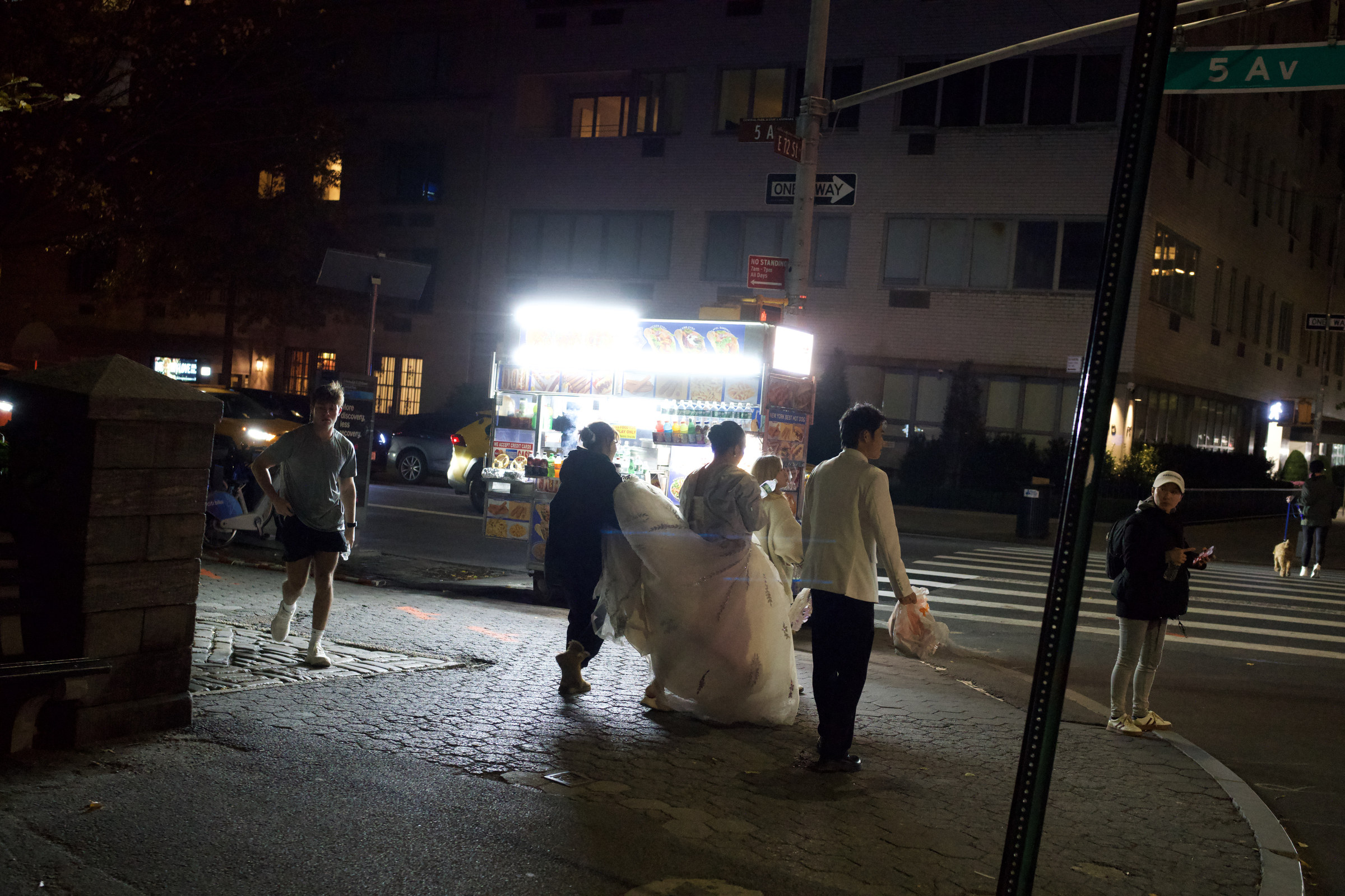 A bride crosses 5th Avenue at night, her white dress billowing as her entourage follows, a glowing food cart and jogger passing in the background