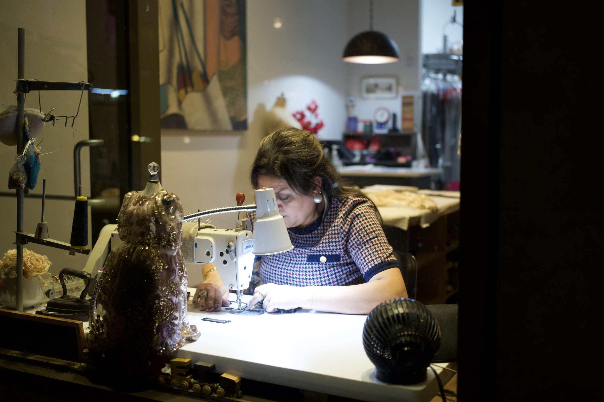 A seamstress hunches over her sewing machine in a shop window at night, an ornate beaded dress form glowing beside her