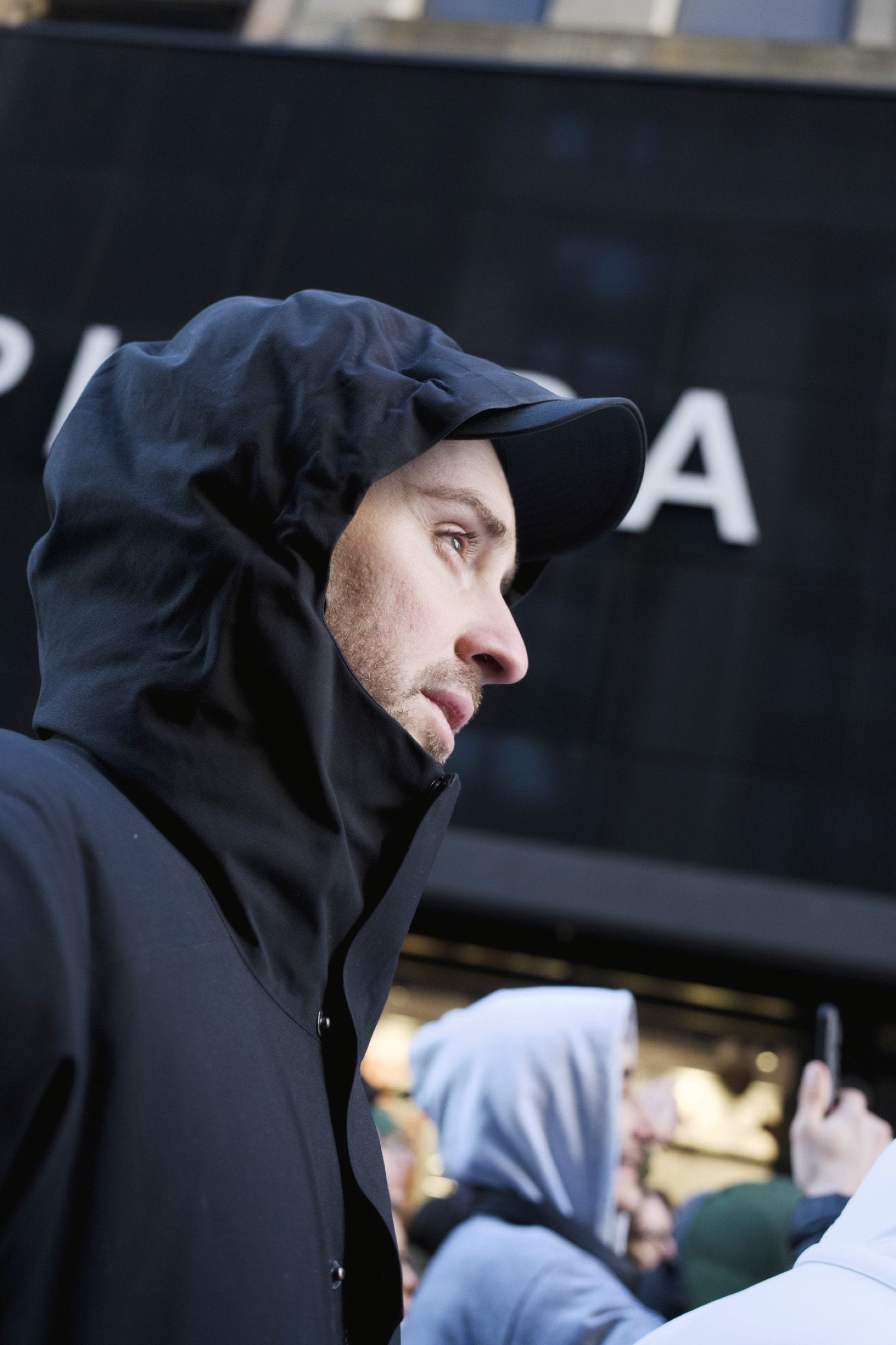 Man in an all-black hooded jacket seen in sharp profile against the PRADA storefront sign, a sculptural silhouette against luxury commerce