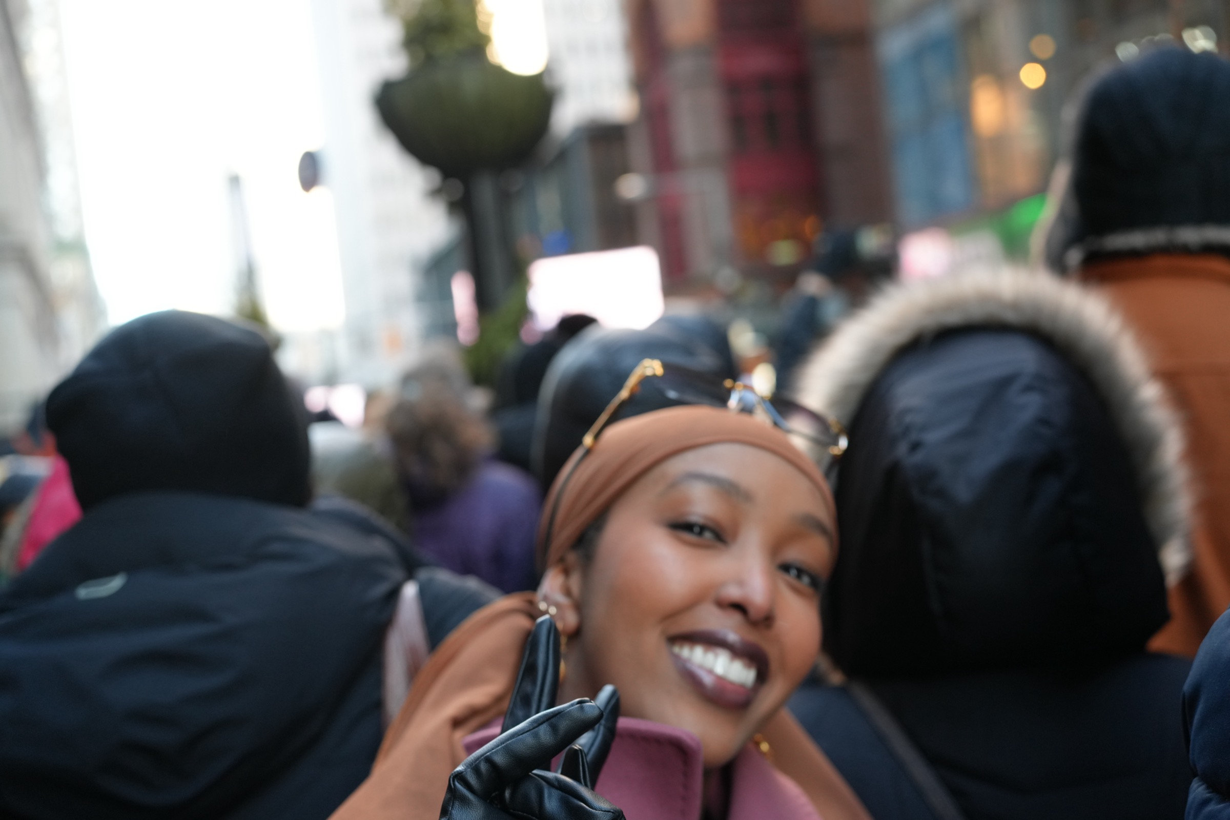 Young woman in a caramel headscarf and leather gloves beams directly into the lens, the crowd dissolving behind her radiant smile