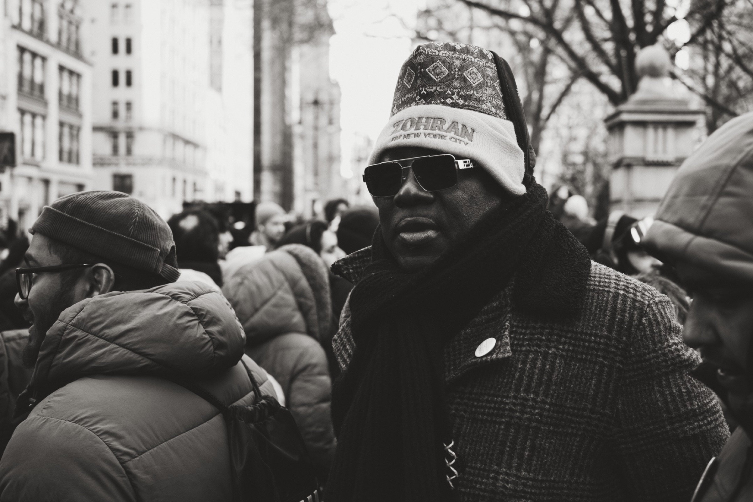 Man in a Zohran for New York City beanie and plaid overcoat stands solid in the inauguration crowd, aviator sunglasses catching the winter light, black and white