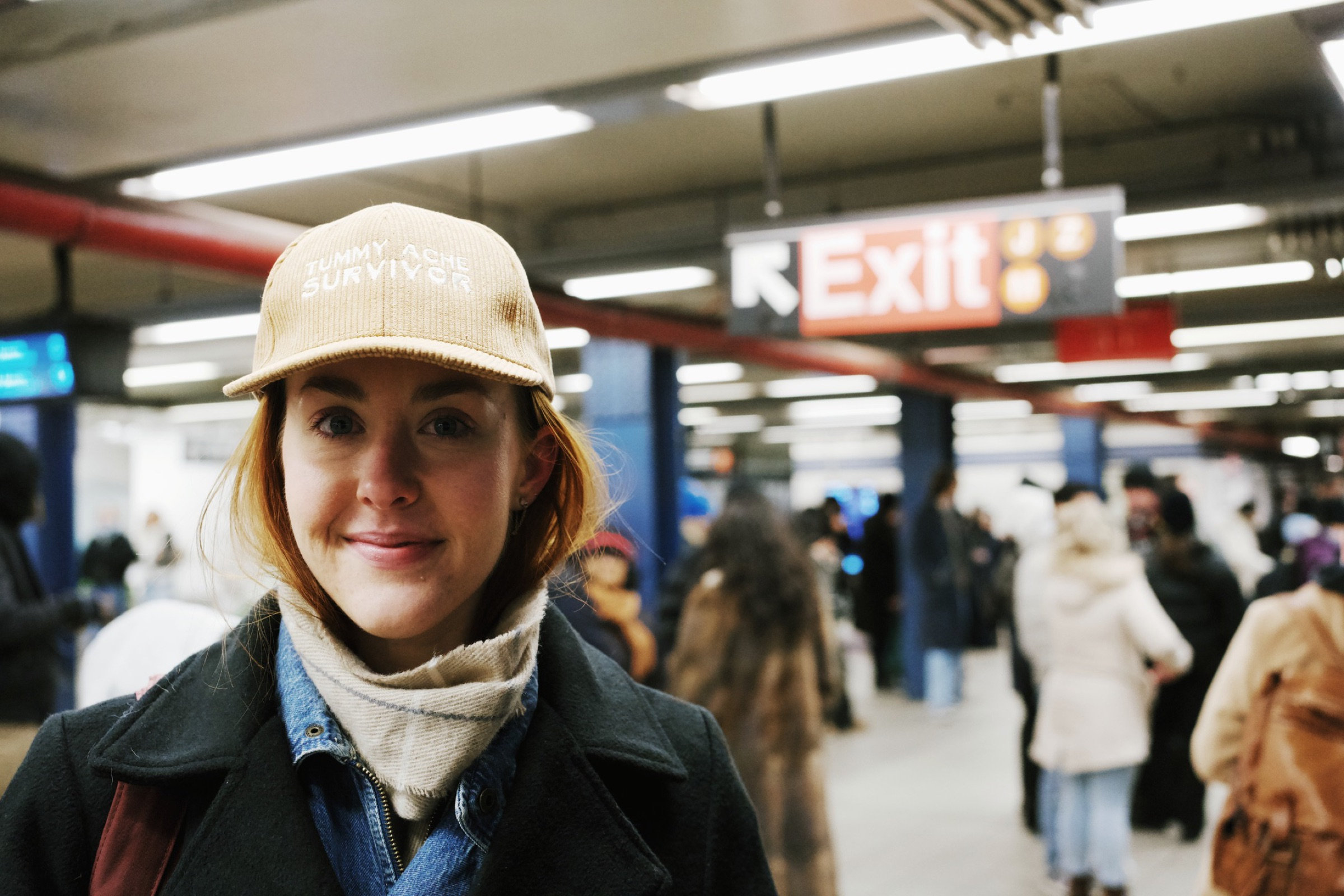 Young woman in a Tummy Ache Survivor corduroy cap smiles gently in the subway, a moment of quiet humor underground