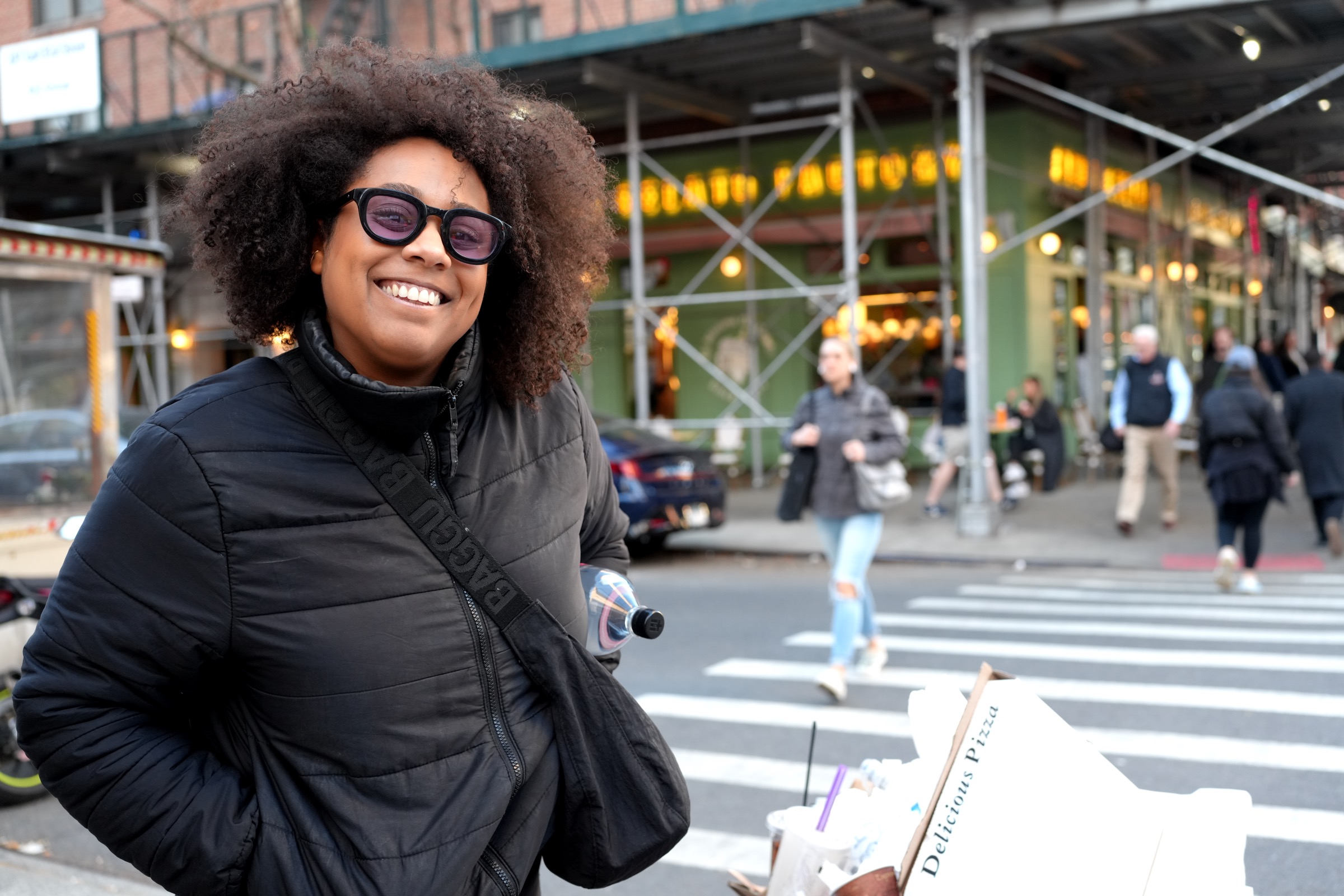 Woman beaming with a Delicious Pizza box under scaffolding at a New York crosswalk, yellow taxi blur behind her