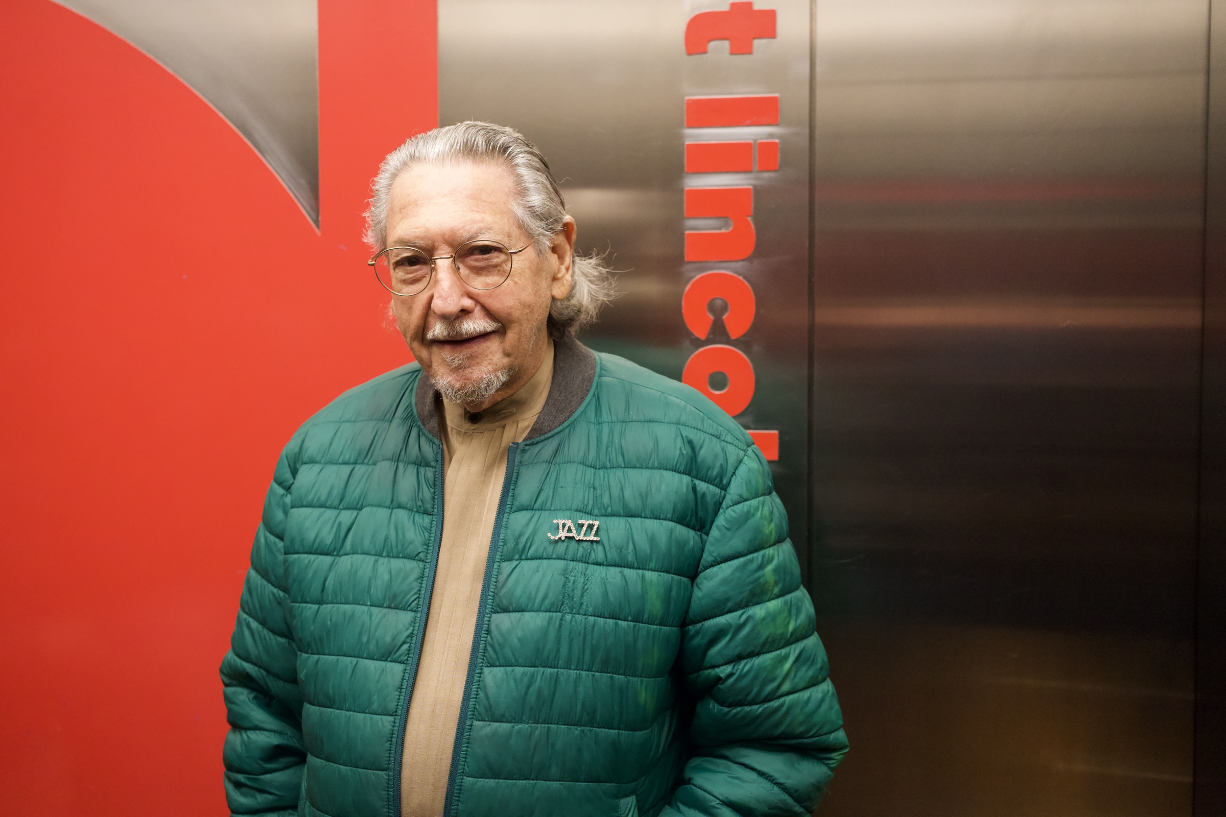 Elderly man with silver hair and glasses in a green puffer jacket with a JAZZ pin smiles warmly in front of red Lincoln Center signage