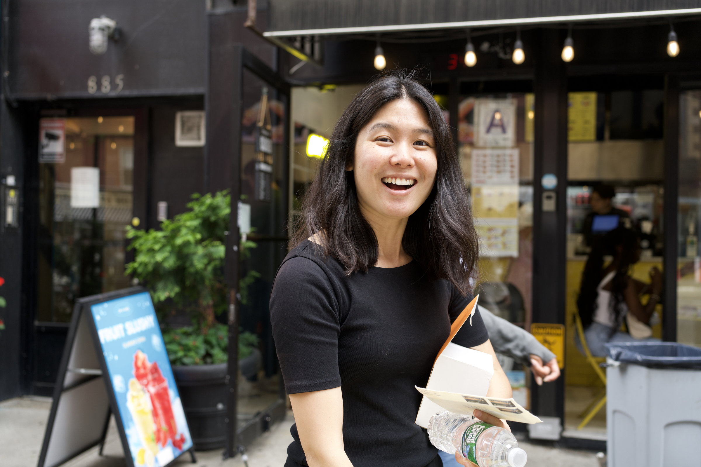 Young woman in a black t-shirt laughs openly on a New York sidewalk, storefronts and string lights behind her
