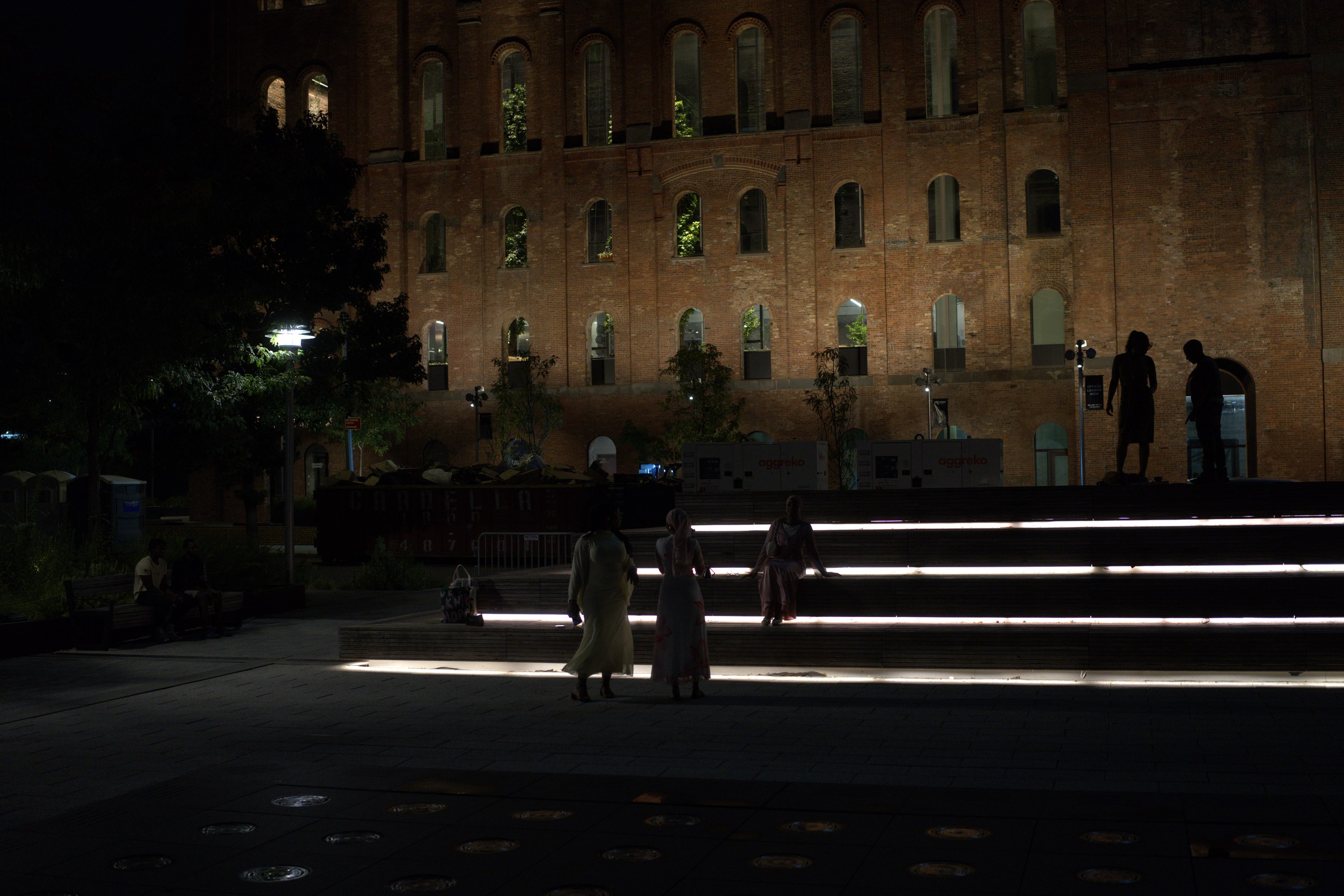 Three women in long dresses approach the LED-lit steps of the Domino Sugar Refinery at night, a silhouetted couple stands above them against the glowing brick facade