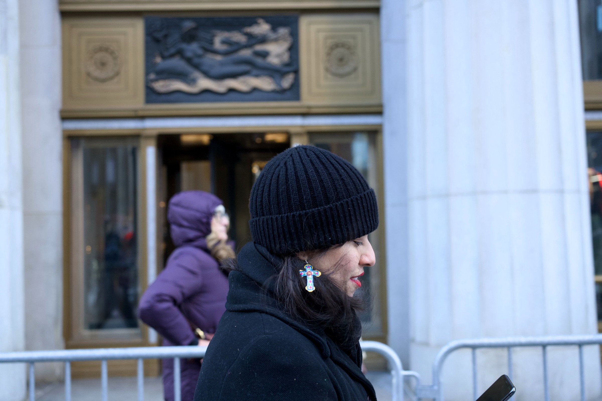 Woman in a black beanie seen in profile, ornate cross earring catching the light, bronze relief sculpture on a grand building facade behind her