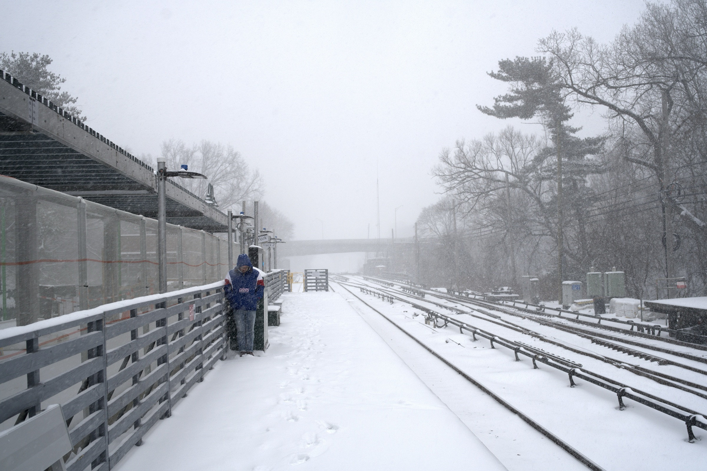 A lone figure in a blue jacket waits on a snow-covered elevated train platform, tracks vanishing into the white void of a blizzard