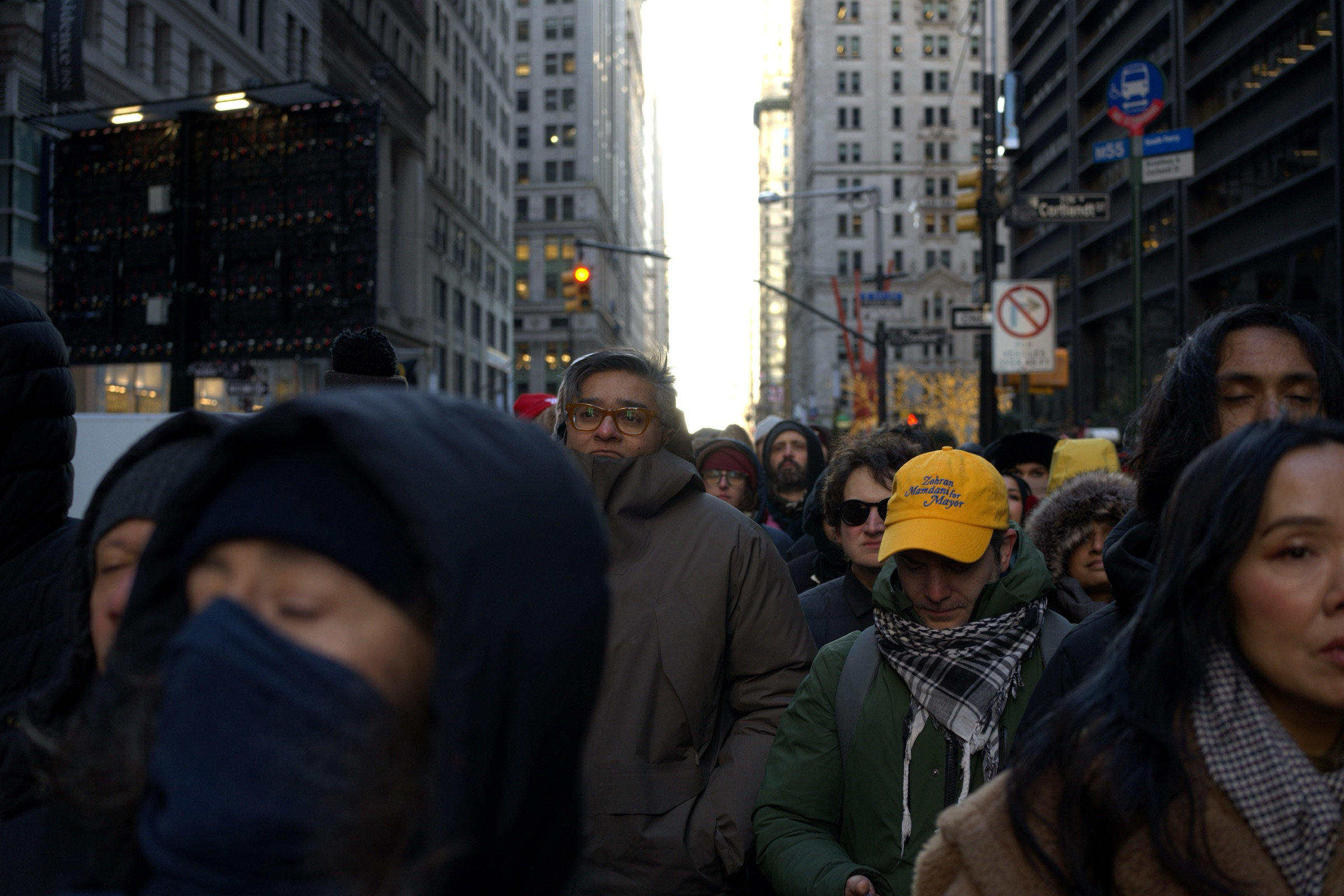 Dense crowd marches through a Lower Manhattan street canyon at golden hour on Inauguration Day, warm backlight pouring down the avenue