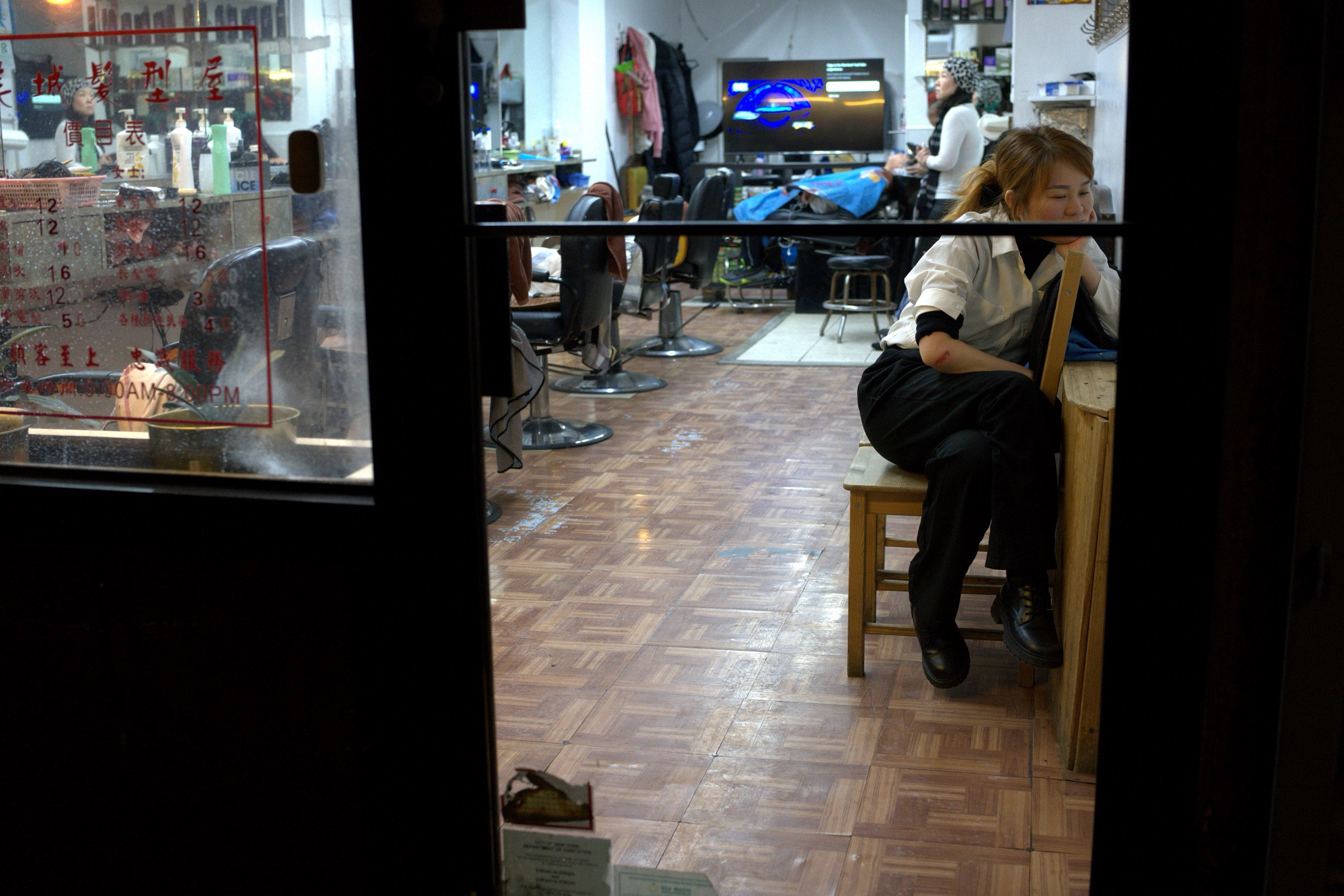 Woman rests in a Chinatown hair salon doorway, eyes closed, hand at her face, framed by the dark door jamb and Chinese signage